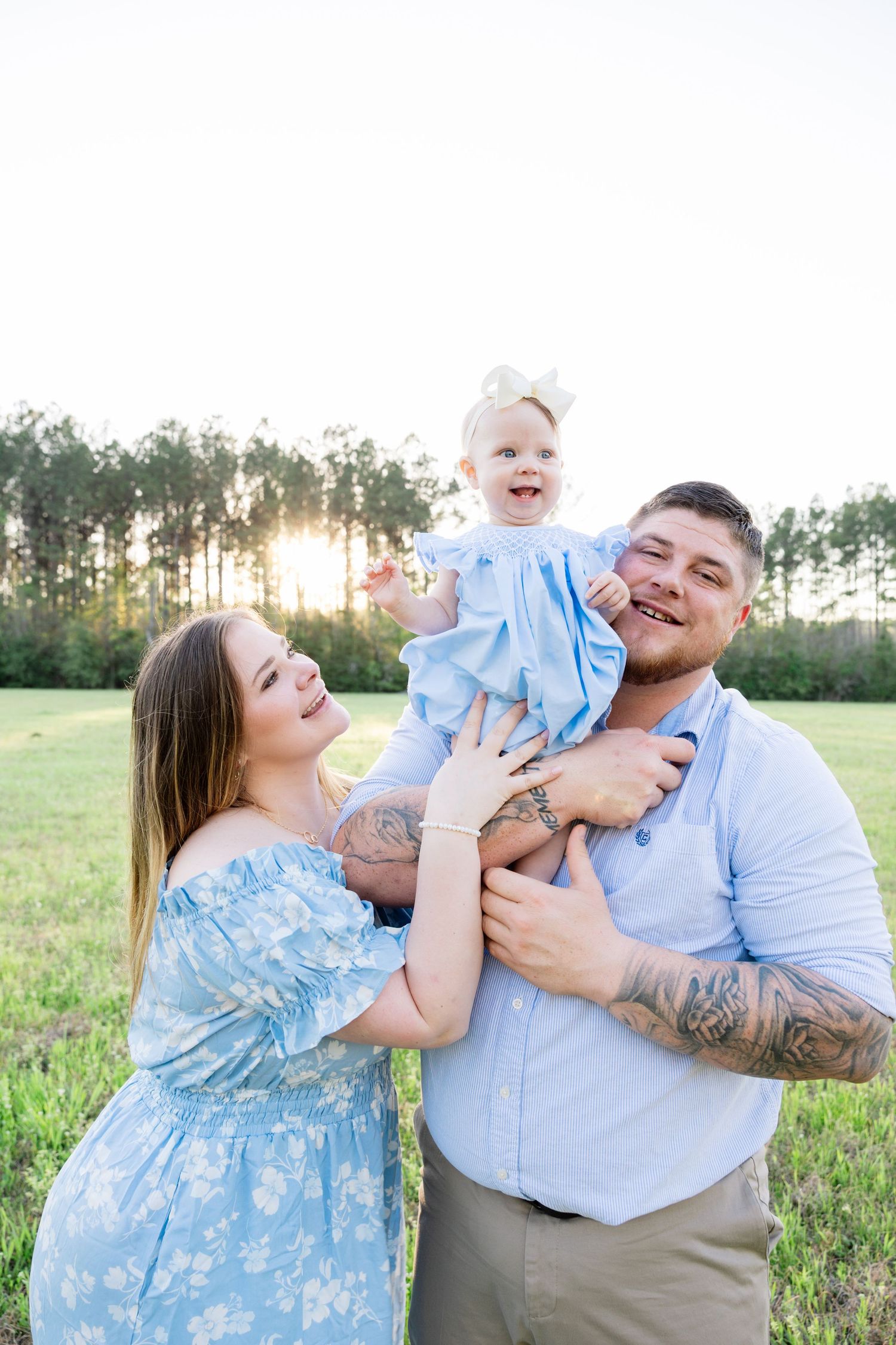 Happy family with tattooed father lifting smiling baby girl in blue dress at golden sunset.