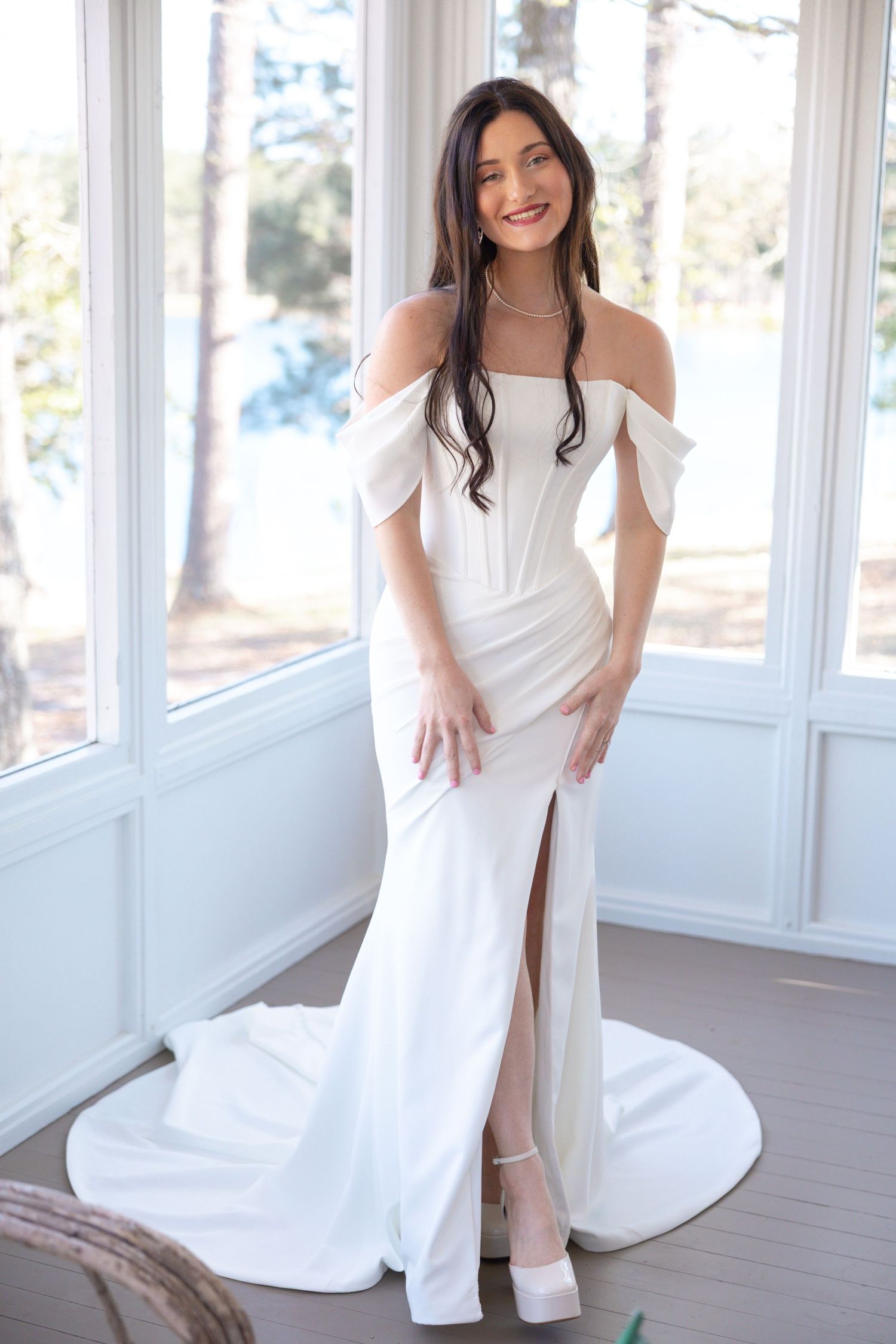 Bride in white off-shoulder fitted wedding gown with front slit posing indoors.