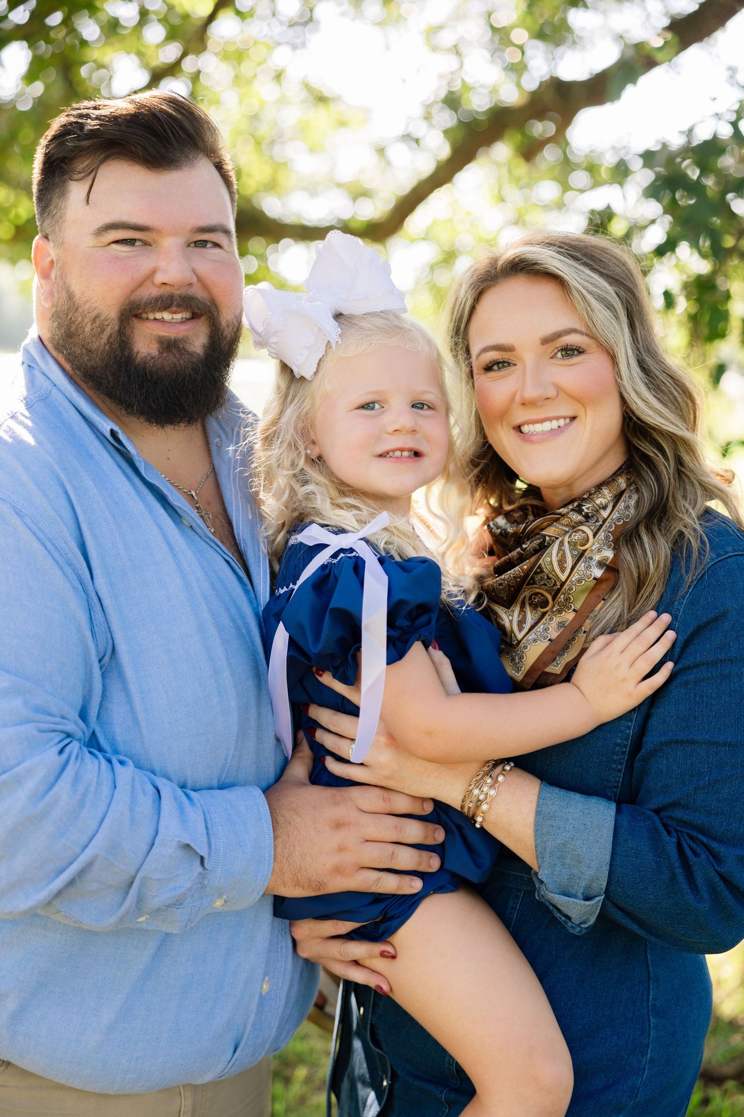Happy family portrait with bearded father, blonde toddler girl with bow, and smiling mother outdoors. Family portraits near Mandeville, LA.