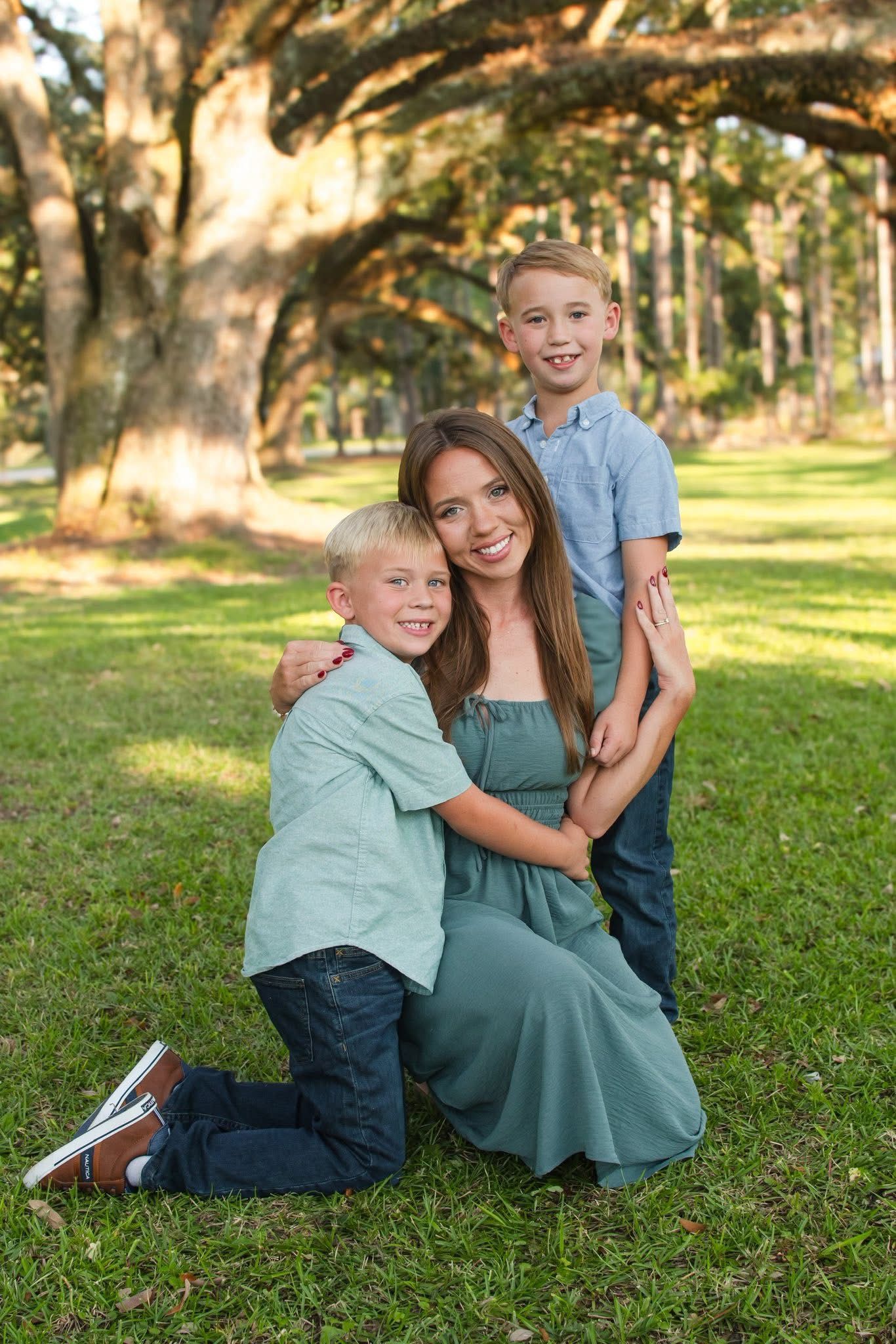 Brunette mother in teal dress kneels on grass hugging two blonde sons under oak trees in park.