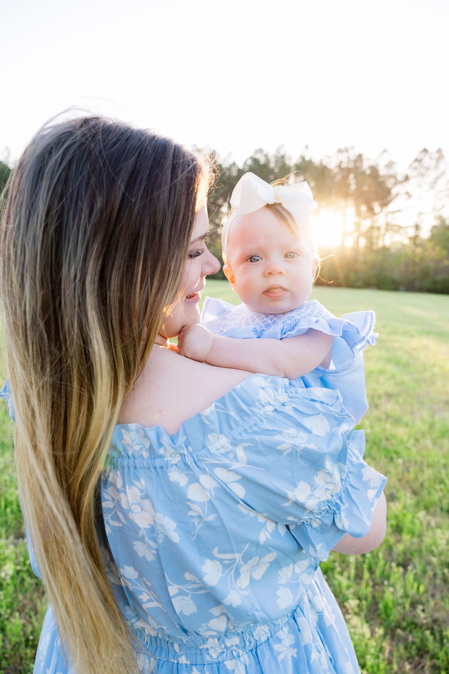 Mother in blue floral dress holds infant with white bow headband at sunny outdoor setting.