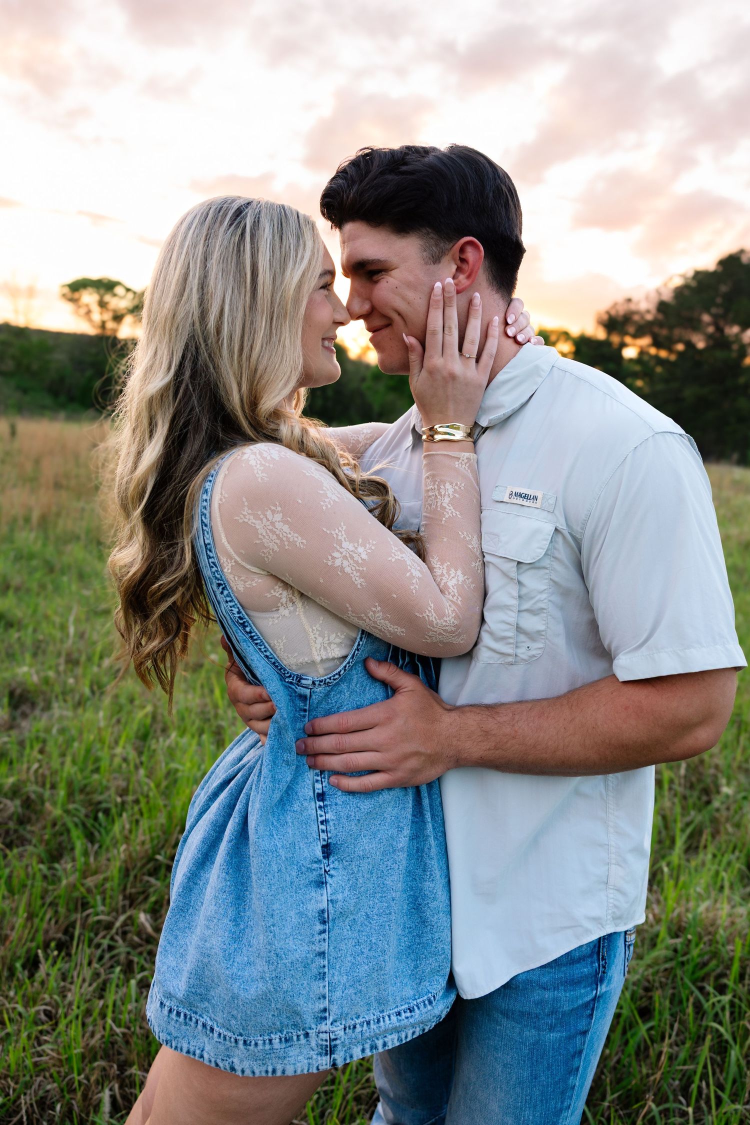 Couple leans in for a kiss in a grassy field during a colorful sunset.