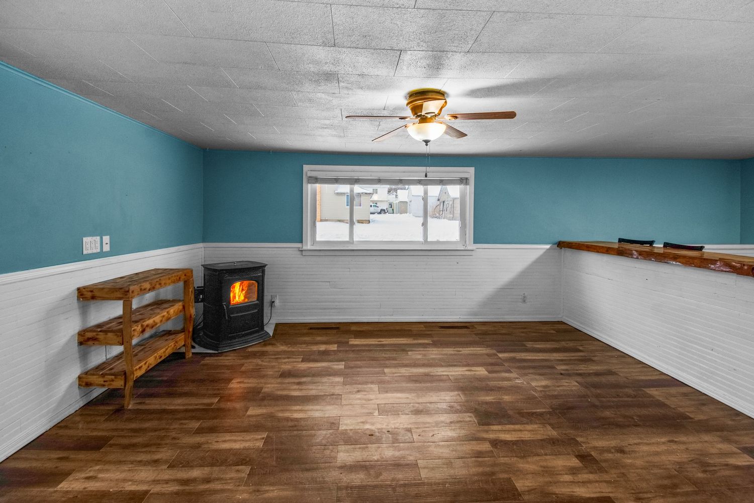 Empty basement room with hardwood floors, turquoise walls, and a corner fireplace beneath a ceiling fan.