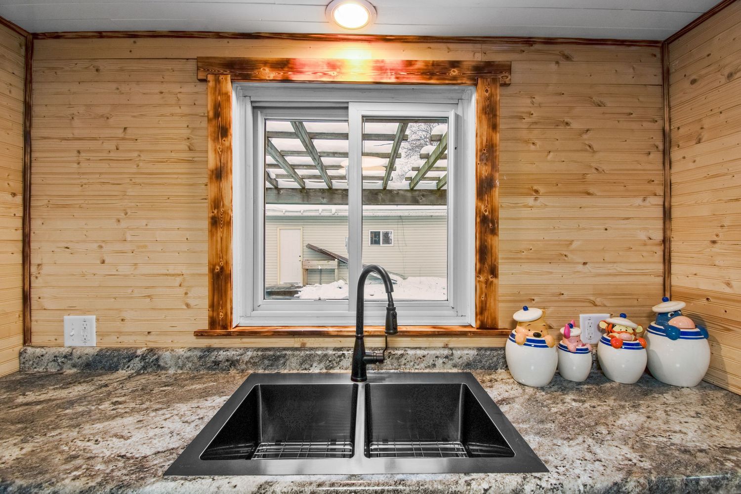 Modern kitchen countertop with dark granite and wood panel backsplash featuring double sink and window.