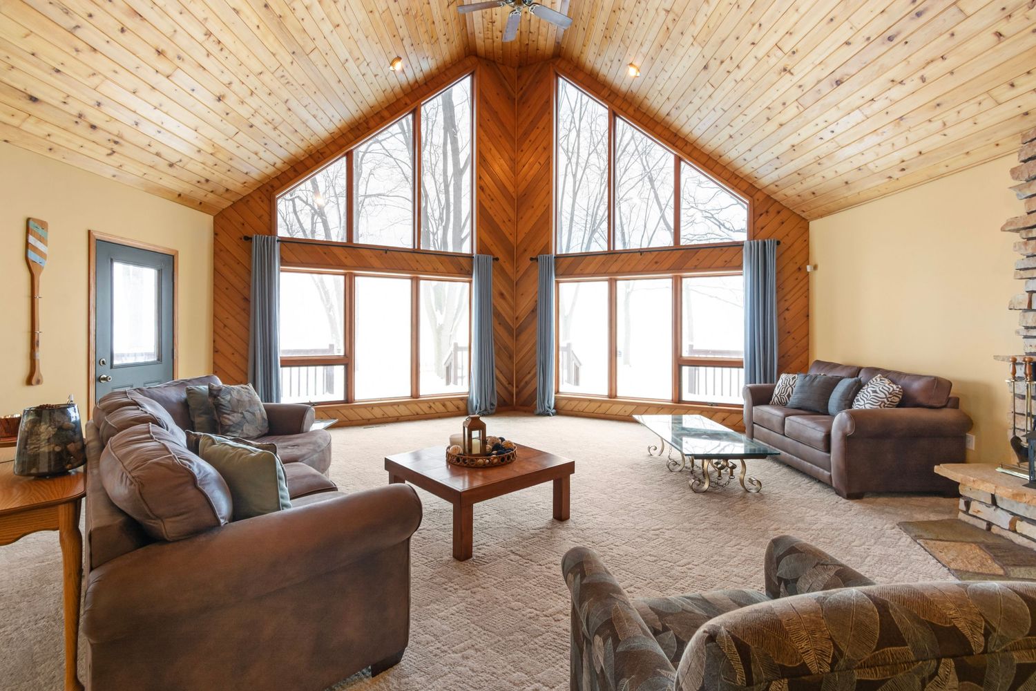 Modern cabin living room with wood-paneled cathedral ceiling, brown furniture, and floor-to-ceiling windows.