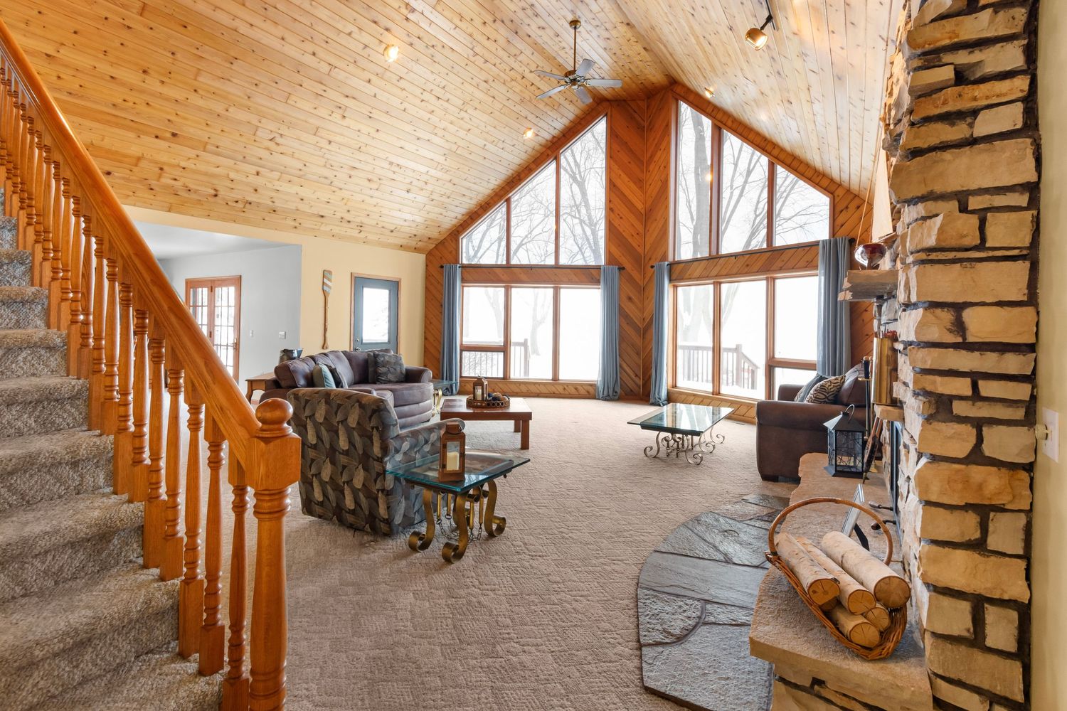 Rustic living room with vaulted wood ceiling, stone fireplace, and large windows showcasing a bright winter landscape.