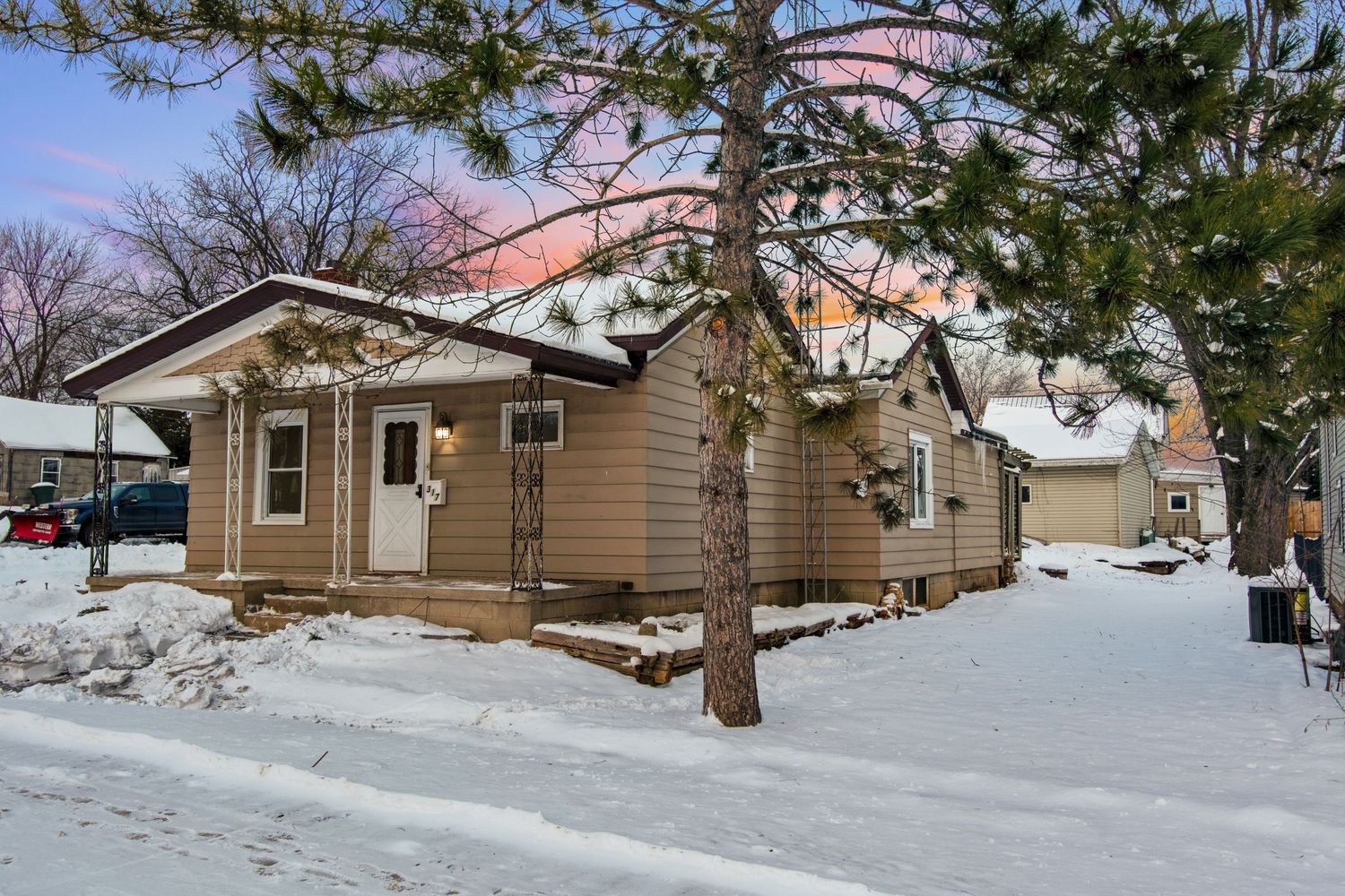 Snow-covered tan bungalow house with pine trees in winter landscape.