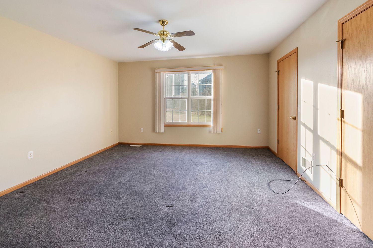 Spacious carpeted living room with beige walls, ceiling fan, and natural light from windows.