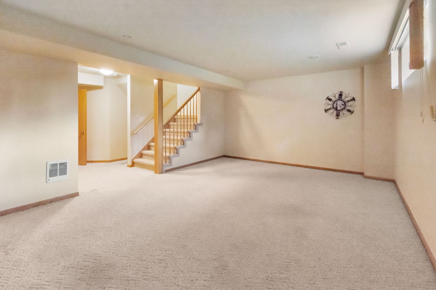 Large basement living area with carpeting, wooden staircase, and decorative wall clock.