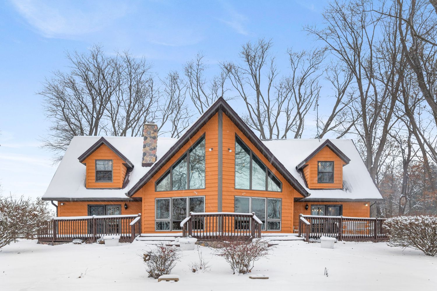 Snow-covered luxury log cabin with large windows, twin dormers, and orange wood exterior in winter setting.