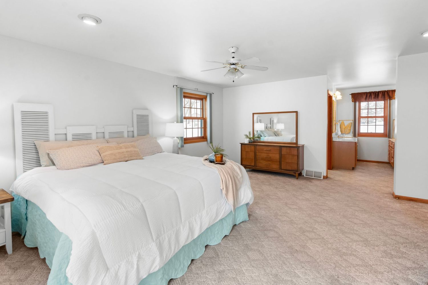 Spacious bedroom with white walls, wood trim windows, beige carpet, and ceiling fan in bright natural light.