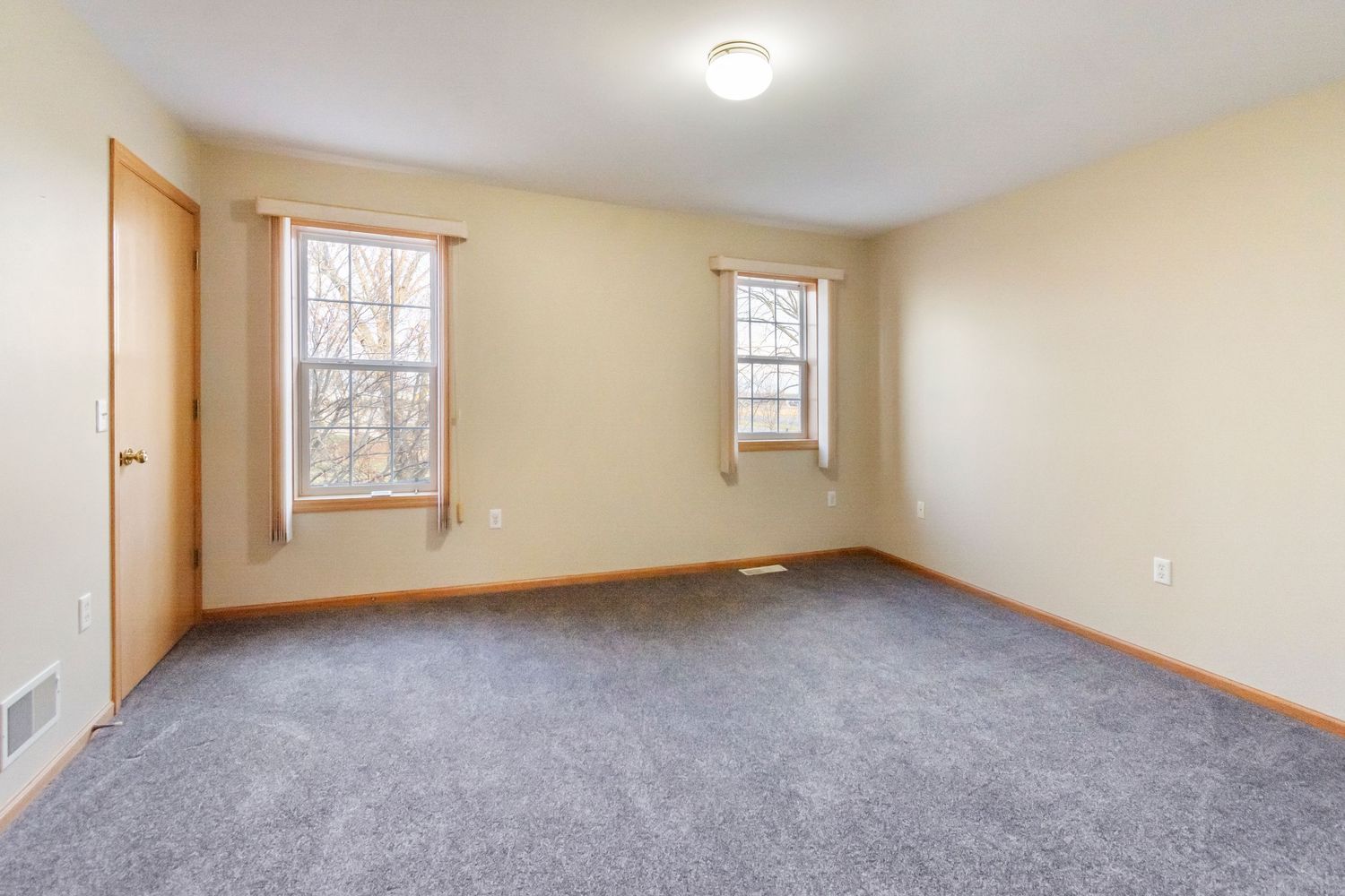 Large unfurnished room with grey carpet, cream colored walls, and multiple windows providing natural light.