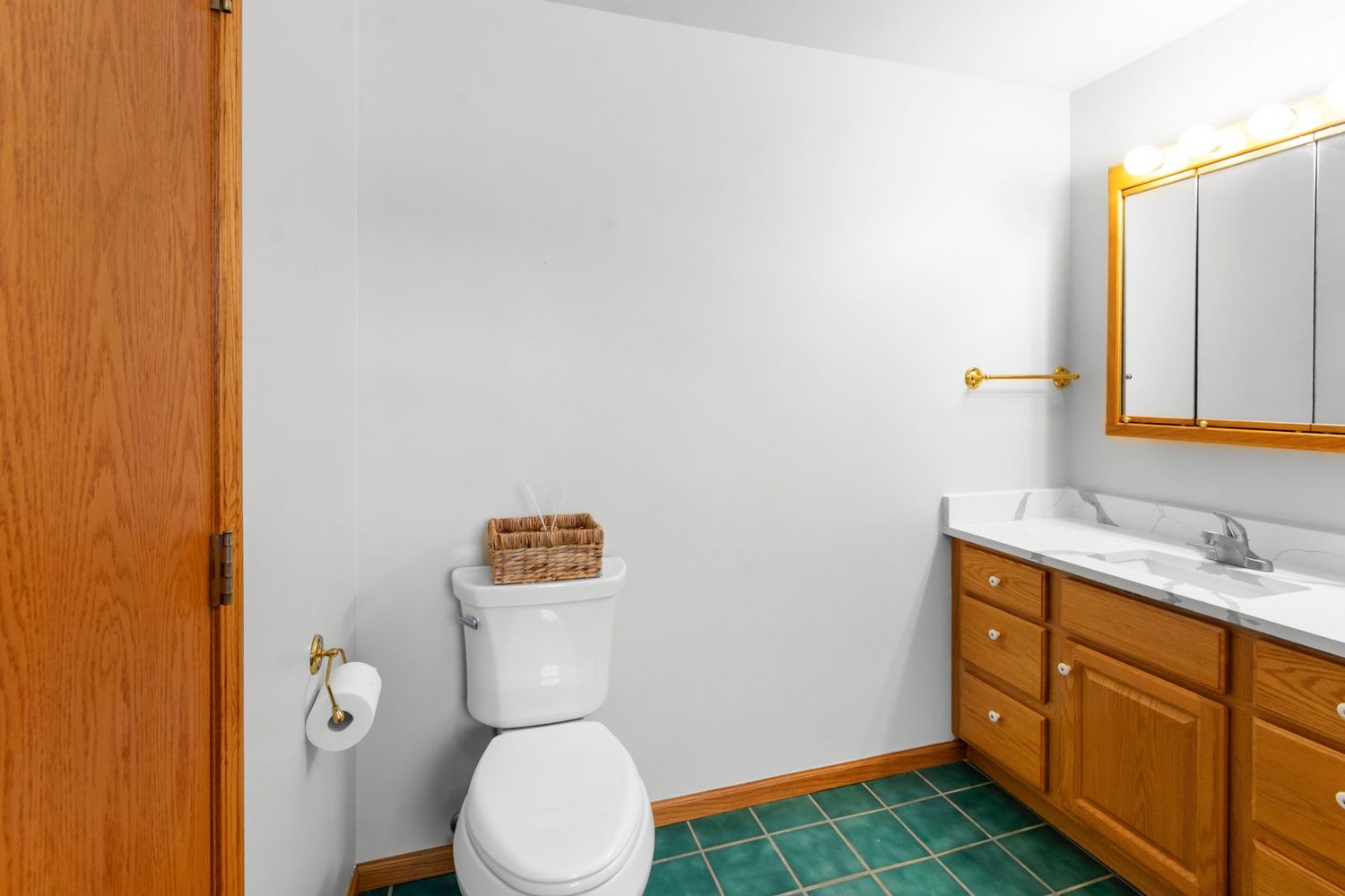 Bright bathroom interior with green tile floor, wooden cabinetry, and white walls featuring a toilet and vanity.