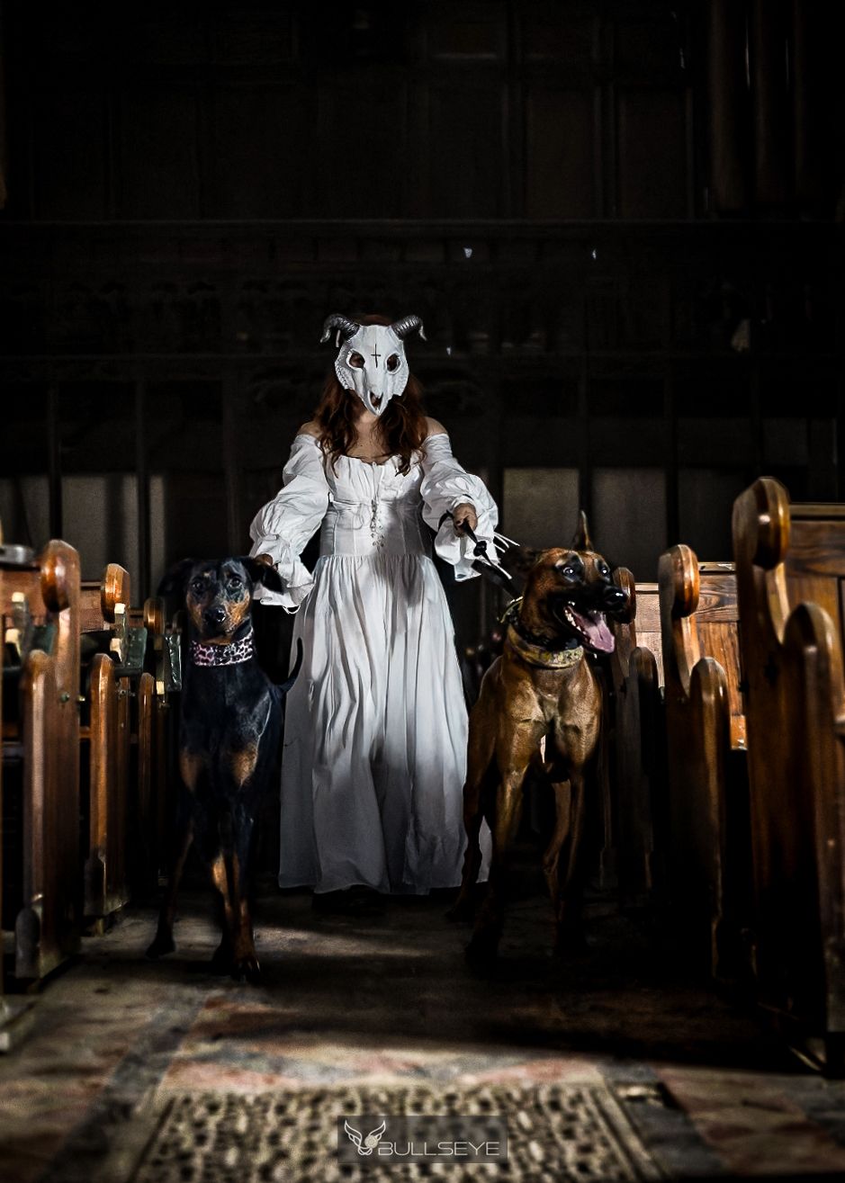 Spooky scene with person in mask and white dress surrounded by dogs in dark room with candles.