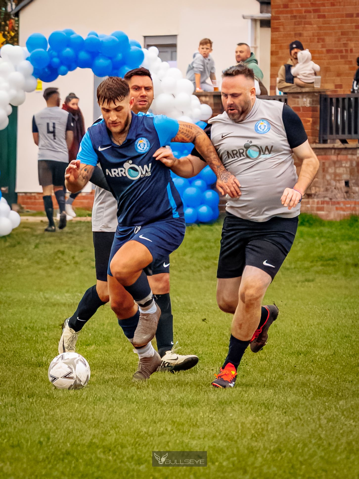Rugby players battle for possession of the ball during an intense match on a grassy field with blue balloons in background.