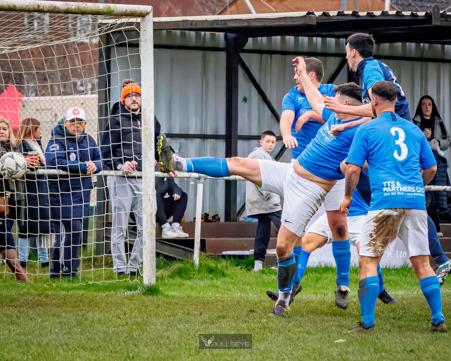 Soccer players in white and blue uniforms compete for the ball near the goal during an intense match.