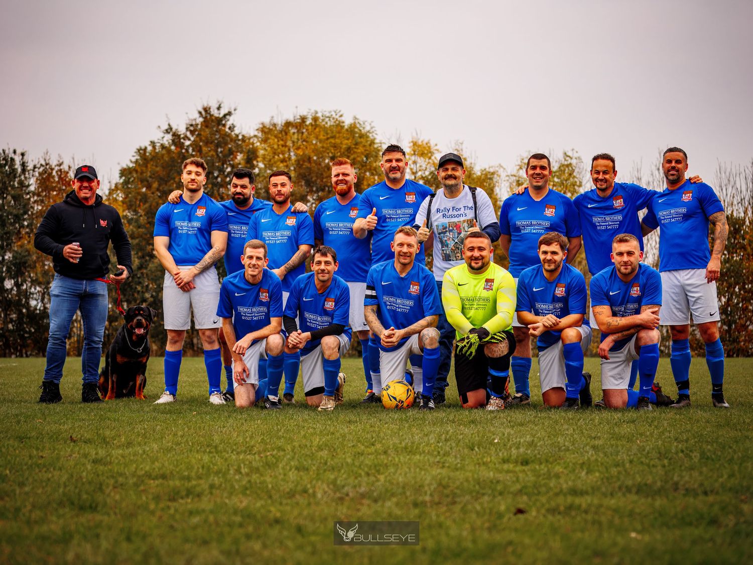 A soccer team in blue jerseys poses together on a grassy field for a group photo at sunset.