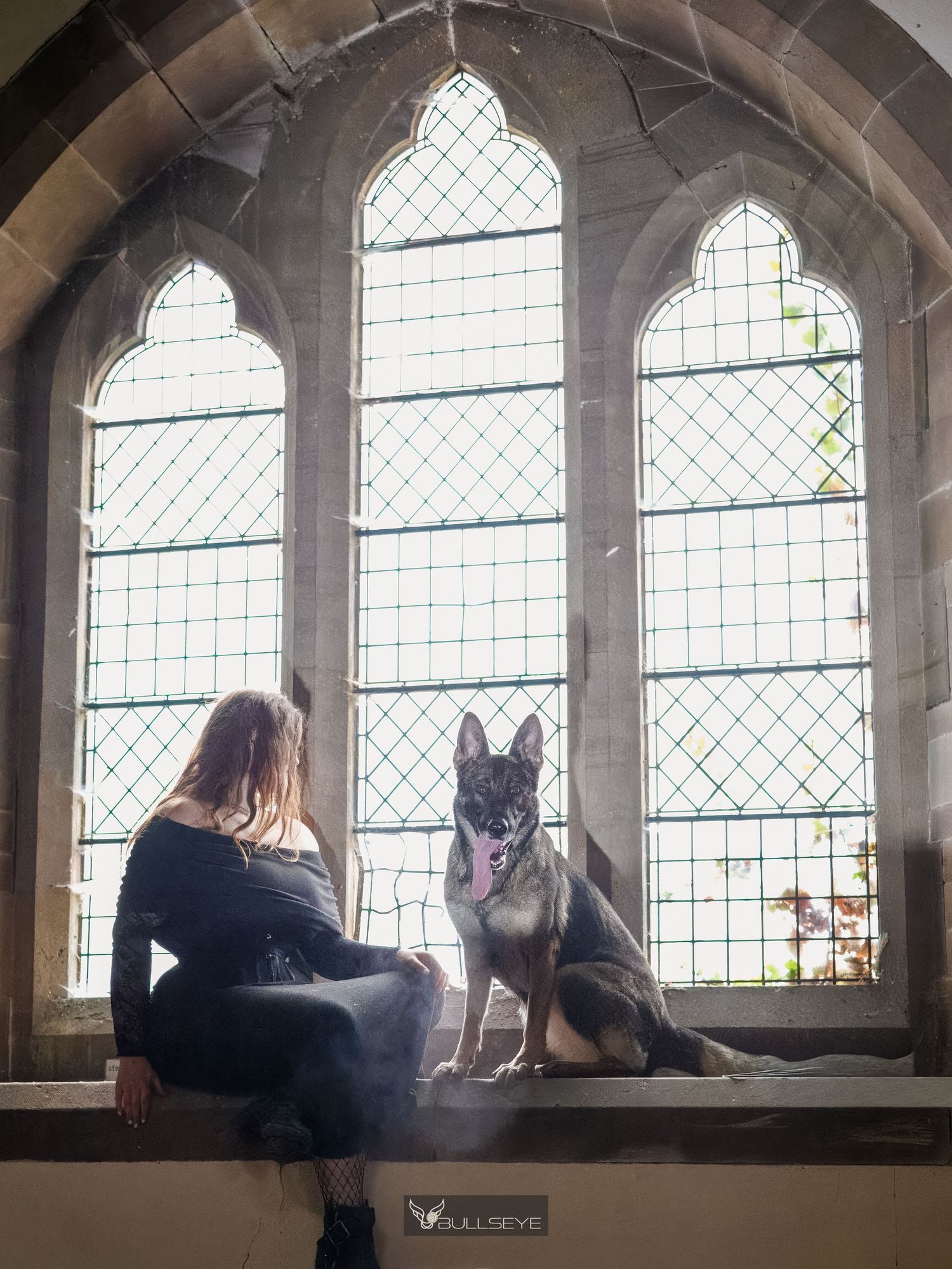 A woman and German Shepherd dog sit on a stone windowsill inside a Gothic church with arched leaded windows.