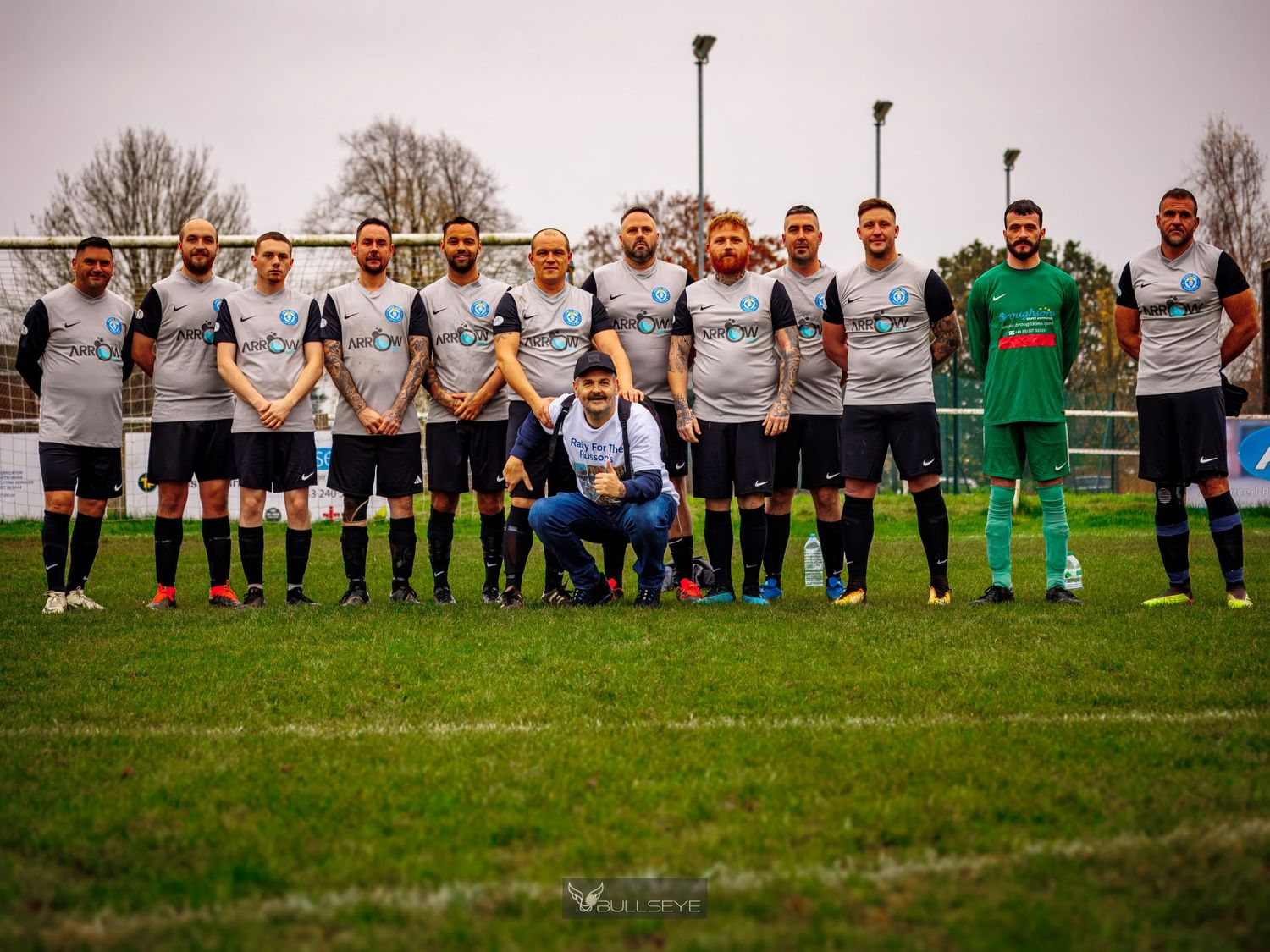 Two soccer teams line up on a grass field before a match with spectators watching from the sidelines.