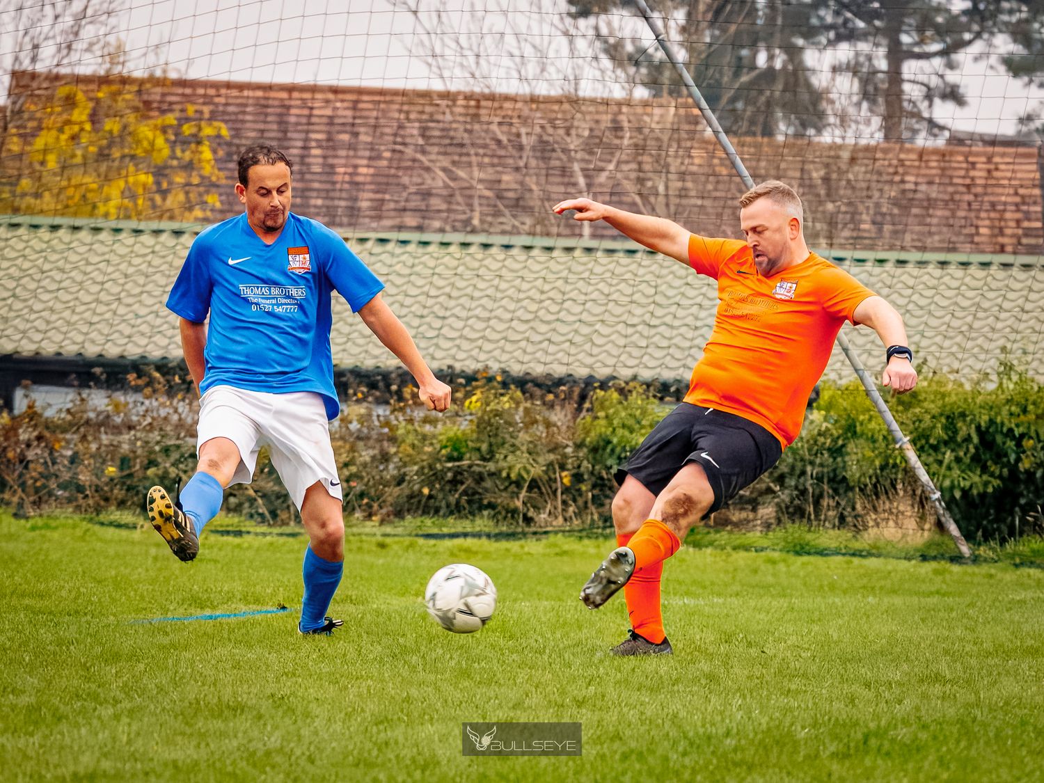Soccer players in orange and blue uniforms compete for the ball on a grass field during an autumn match.