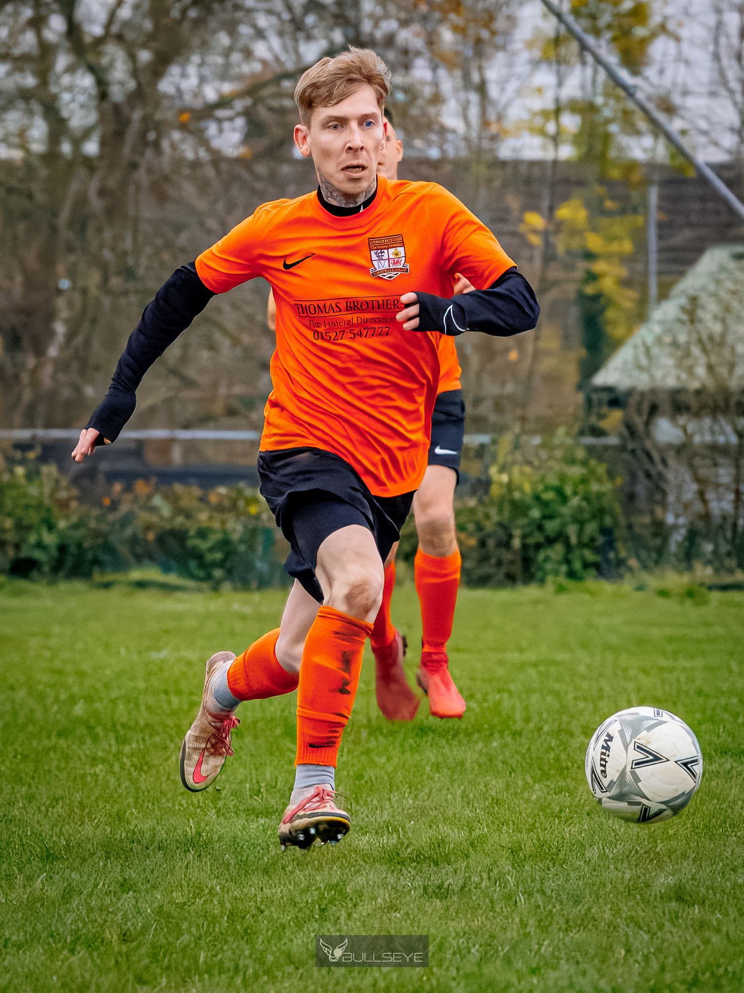 Soccer player in orange jersey and black shorts leaping to kick ball during outdoor match on grass field.