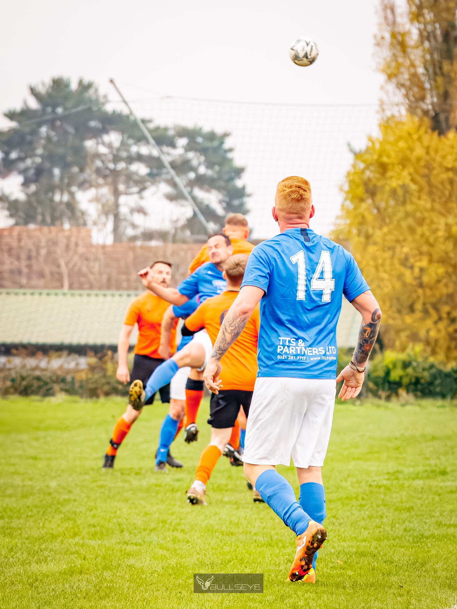 Amateur soccer match action showing players in orange and blue jerseys battling for possession on a fall day.