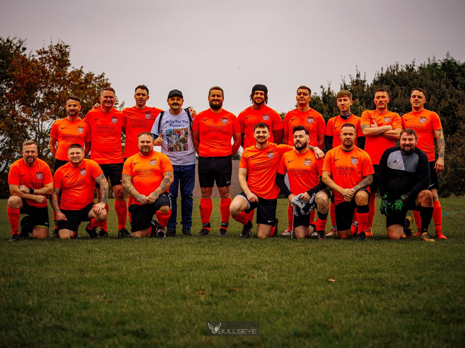 Soccer team wearing bright orange jerseys poses together on grass field for team photo at sunset.