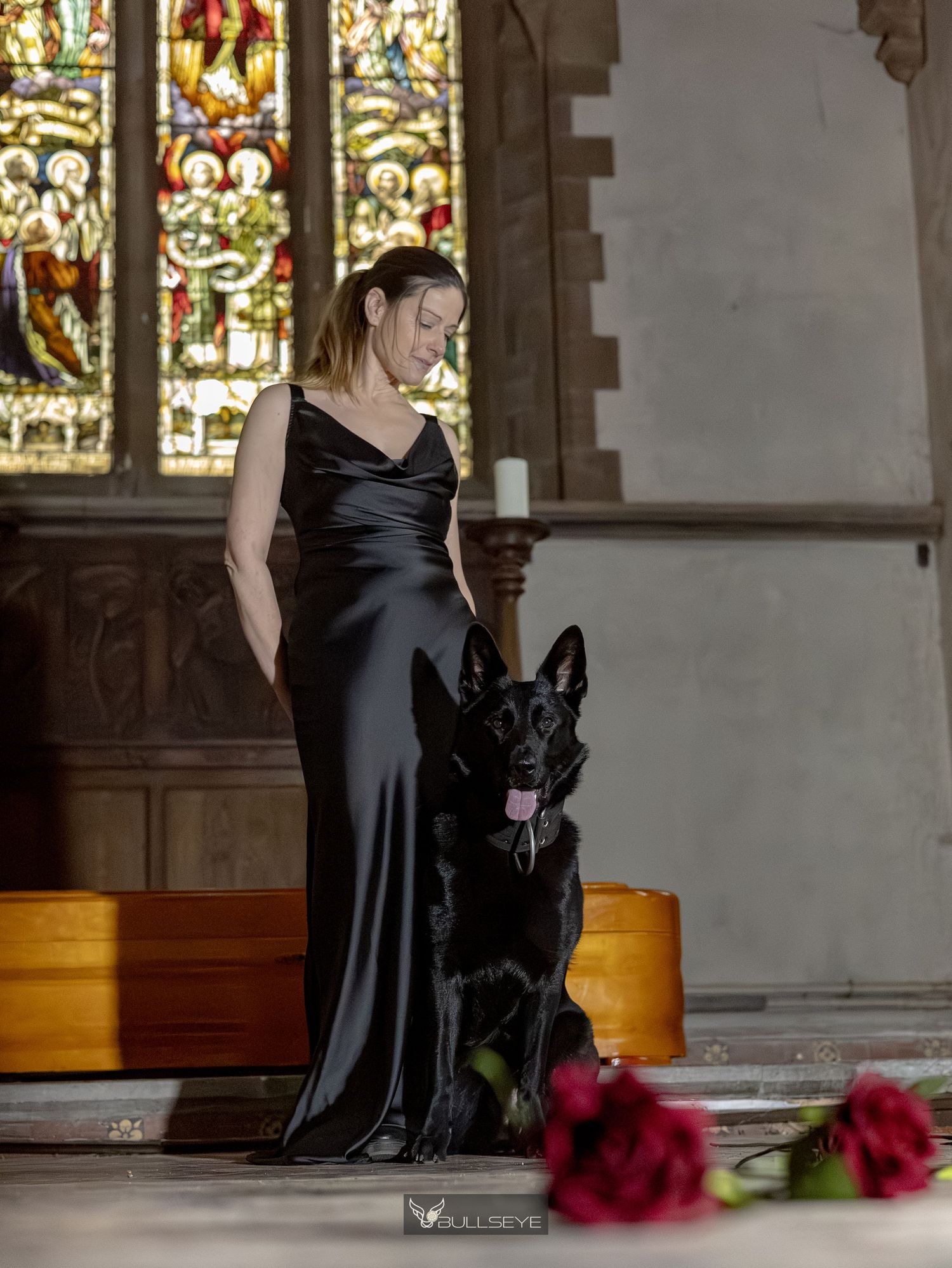 Woman in black satin gown stands with black German Shepherd in church with stained glass windows and red roses.