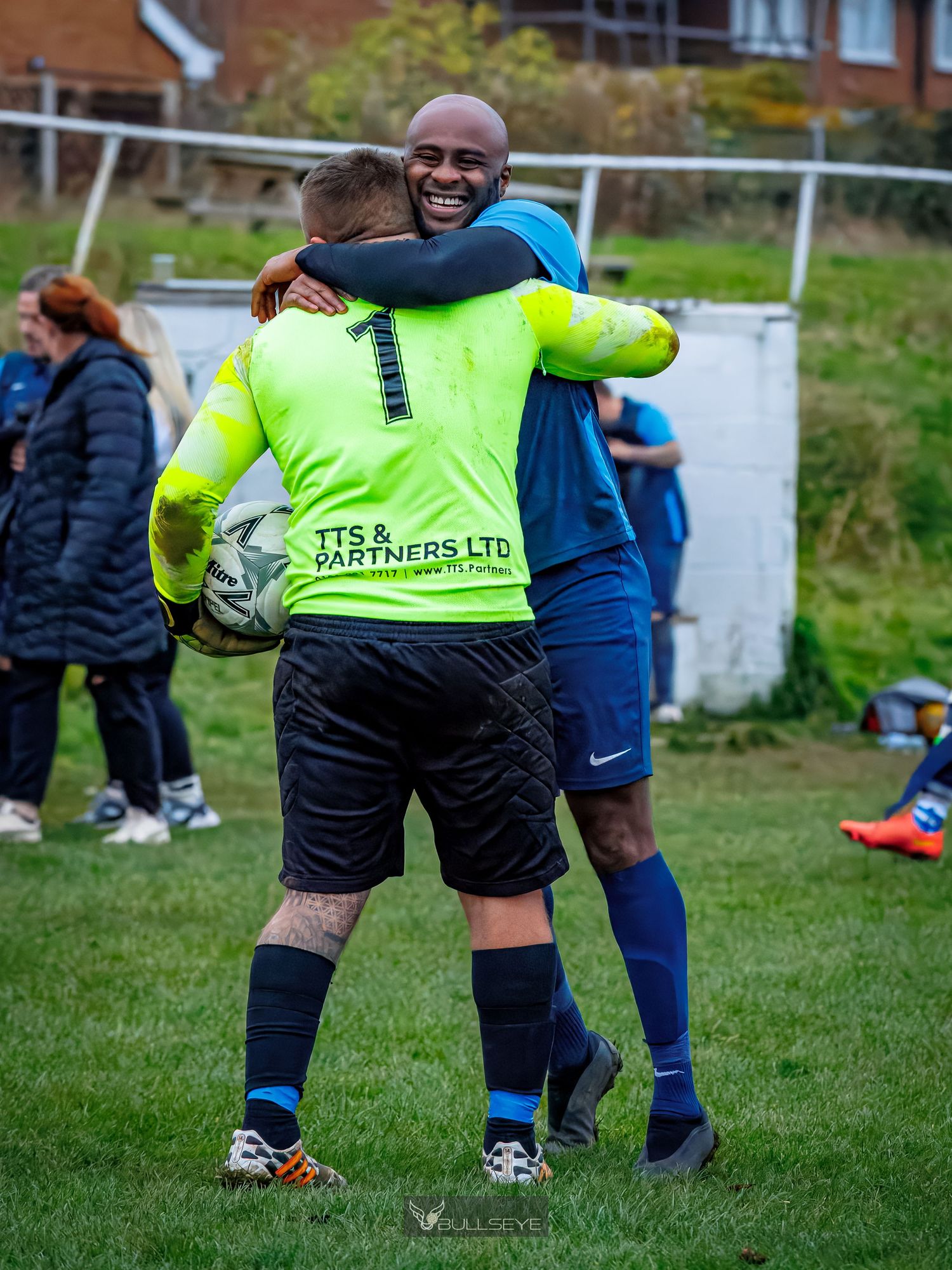 Soccer players share a joyful embrace on the field with one wearing a bright yellow goalkeeper jersey and black shorts.
