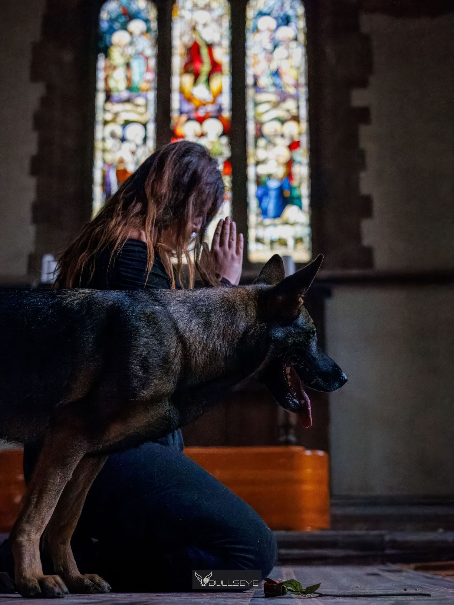 Woman kneeling in prayer with hands clasped beside a German Shepherd dog inside a church with stained glass windows.