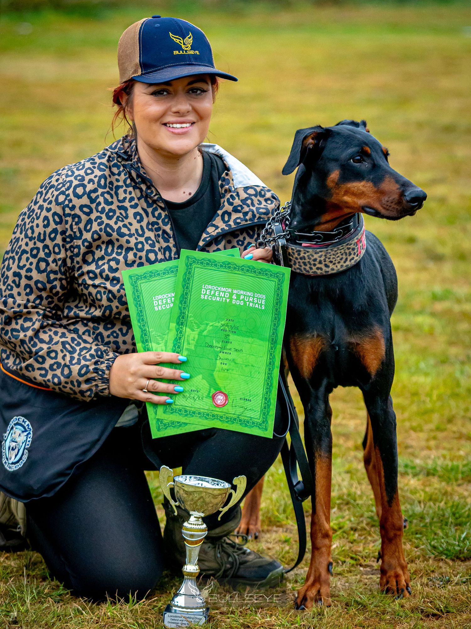 Doberman and handler celebrate with trophy and certificates after competition win.