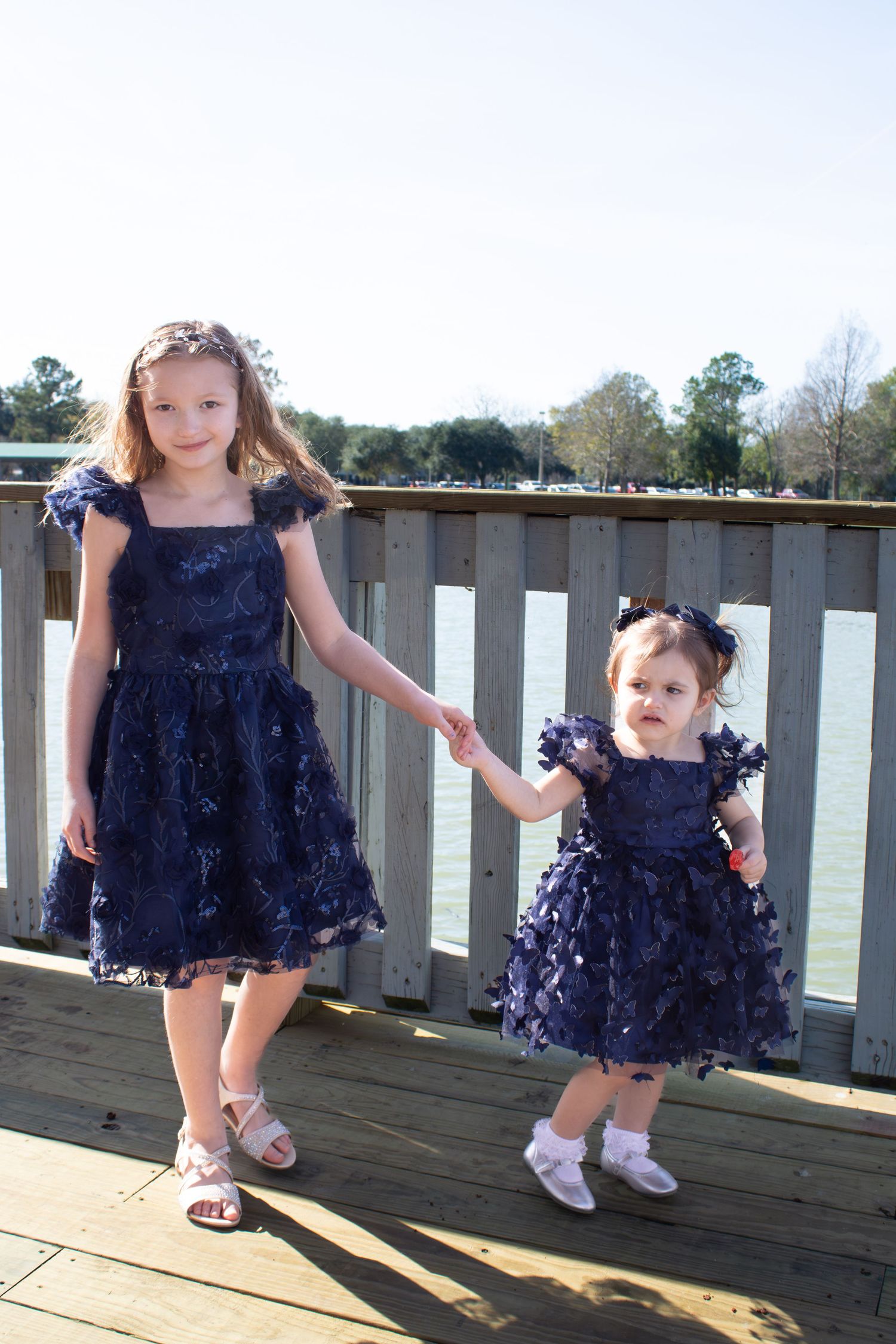 Two children in matching navy blue dresses hold hands on wooden deck.