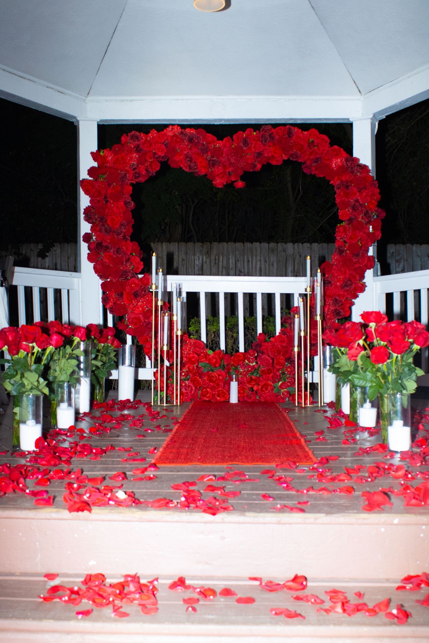 A romantic red rose heart arch with scattered petals decorates a white gazebo.
