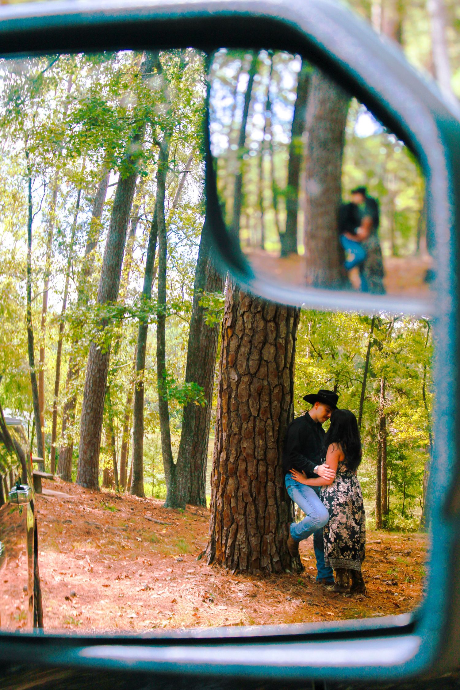 View through car side mirror of a wooded forest path with tall pine trees.