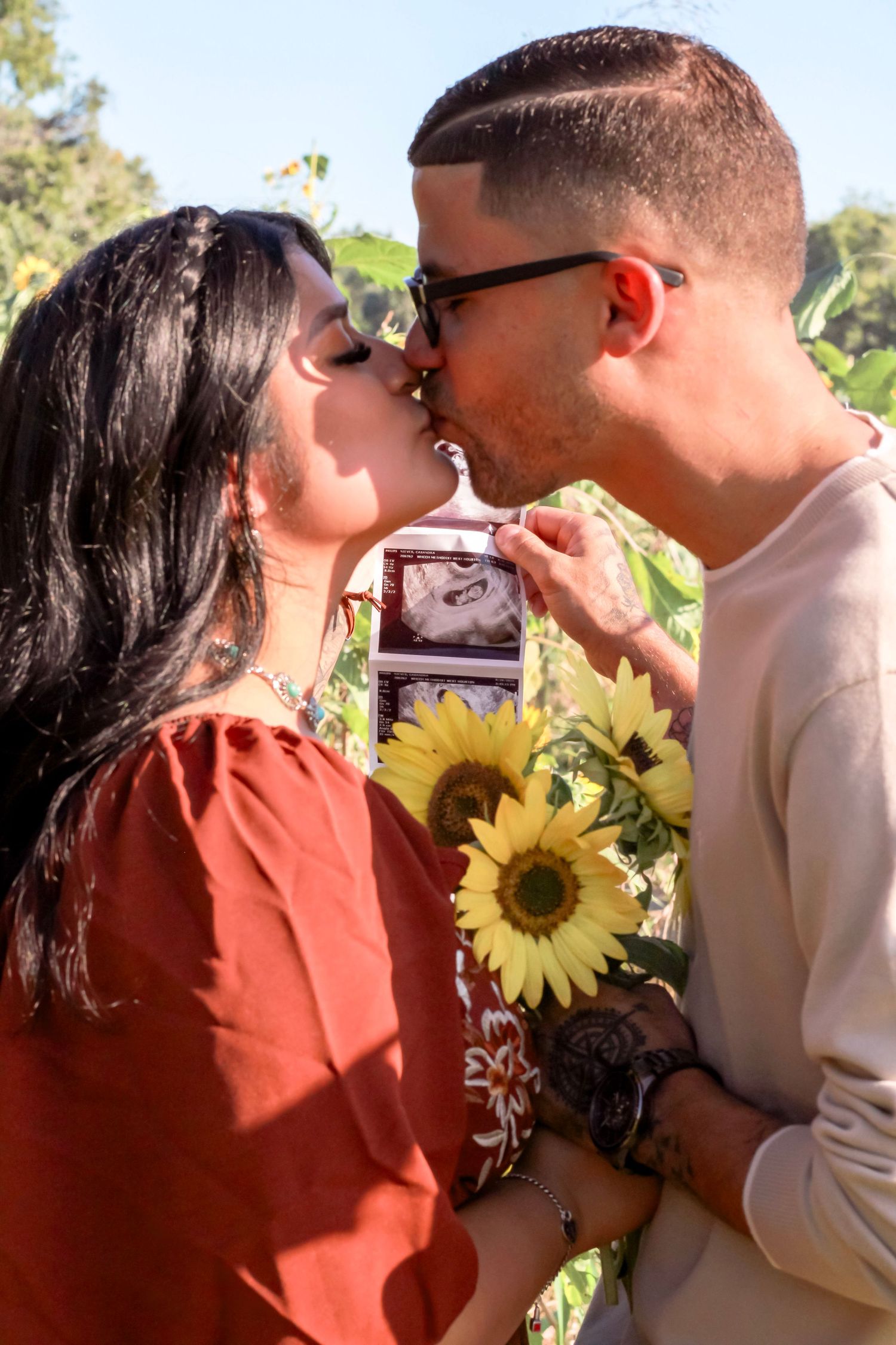 A romantic couple shares a moment with sunflowers and ultrasound photos in sunlight.