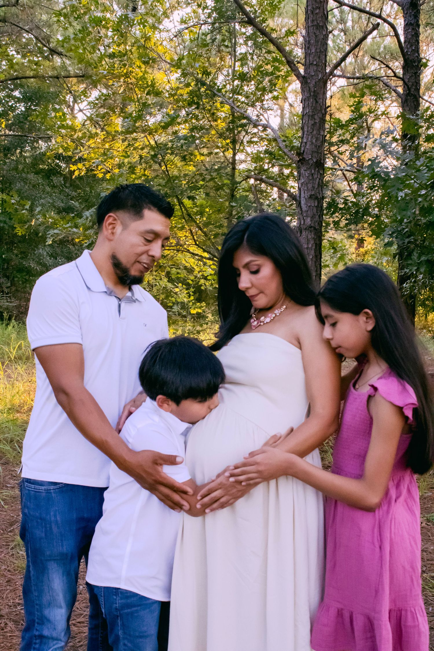 Family gathered for maternity photos in woodland setting wearing coordinated outfits.