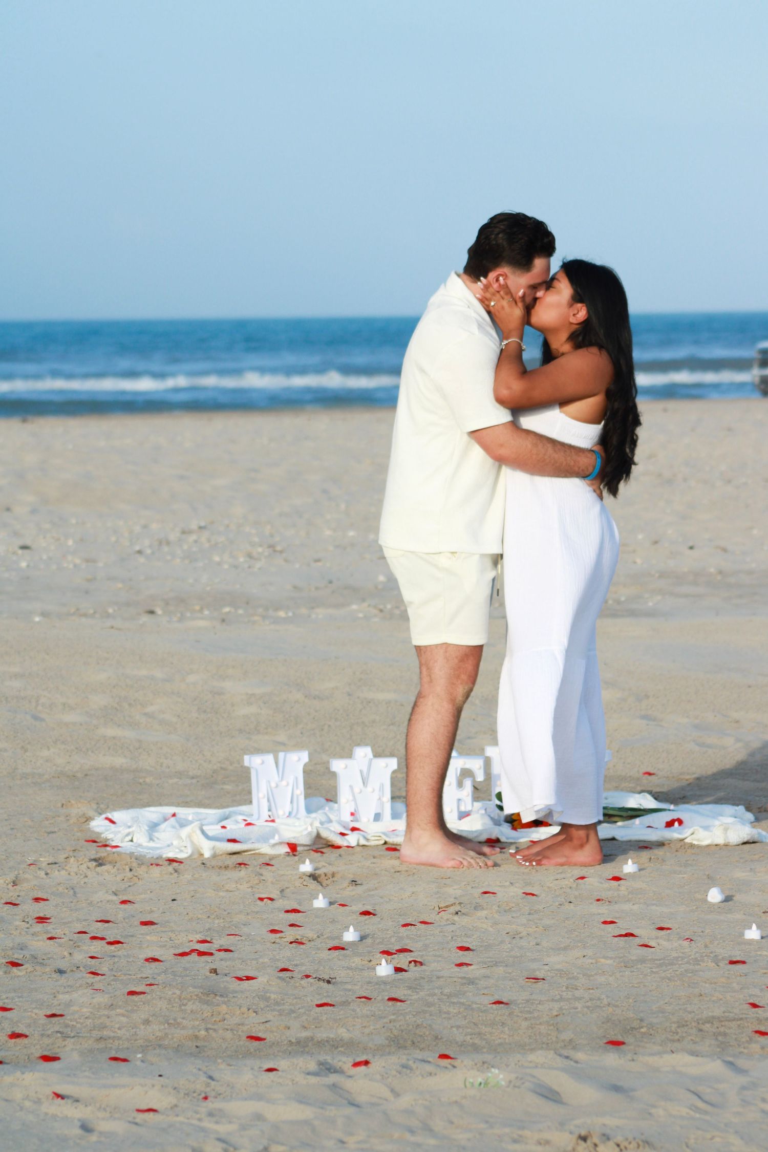 Romantic beach scene with couple in white outfits surrounded by rose petals and decorative letters.