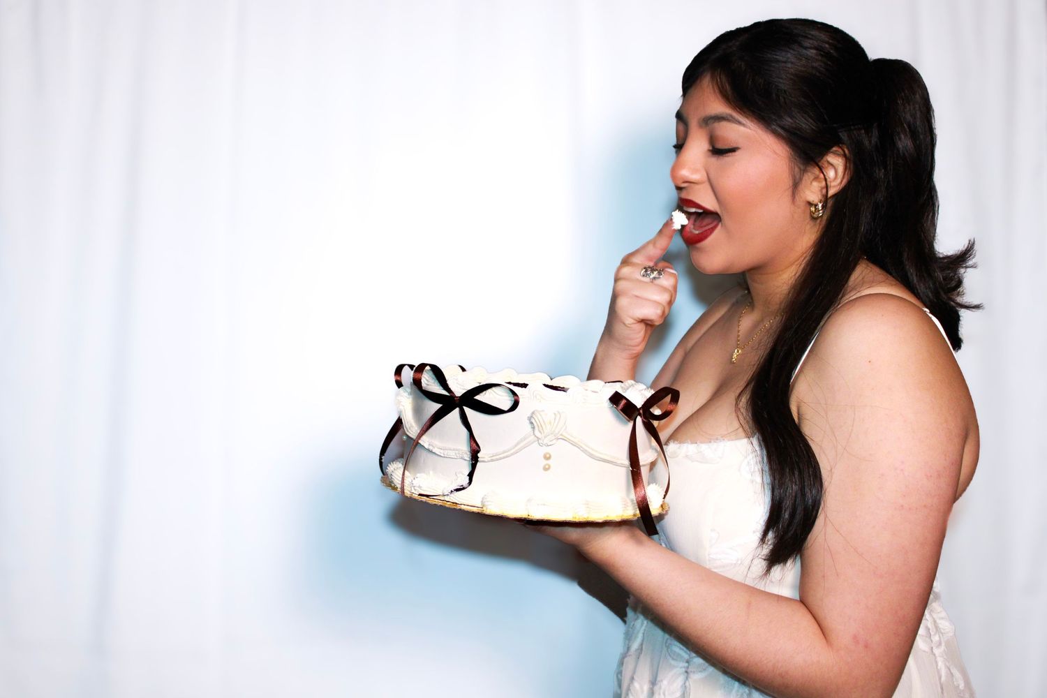 A person in white dress poses playfully with cake against a light background.