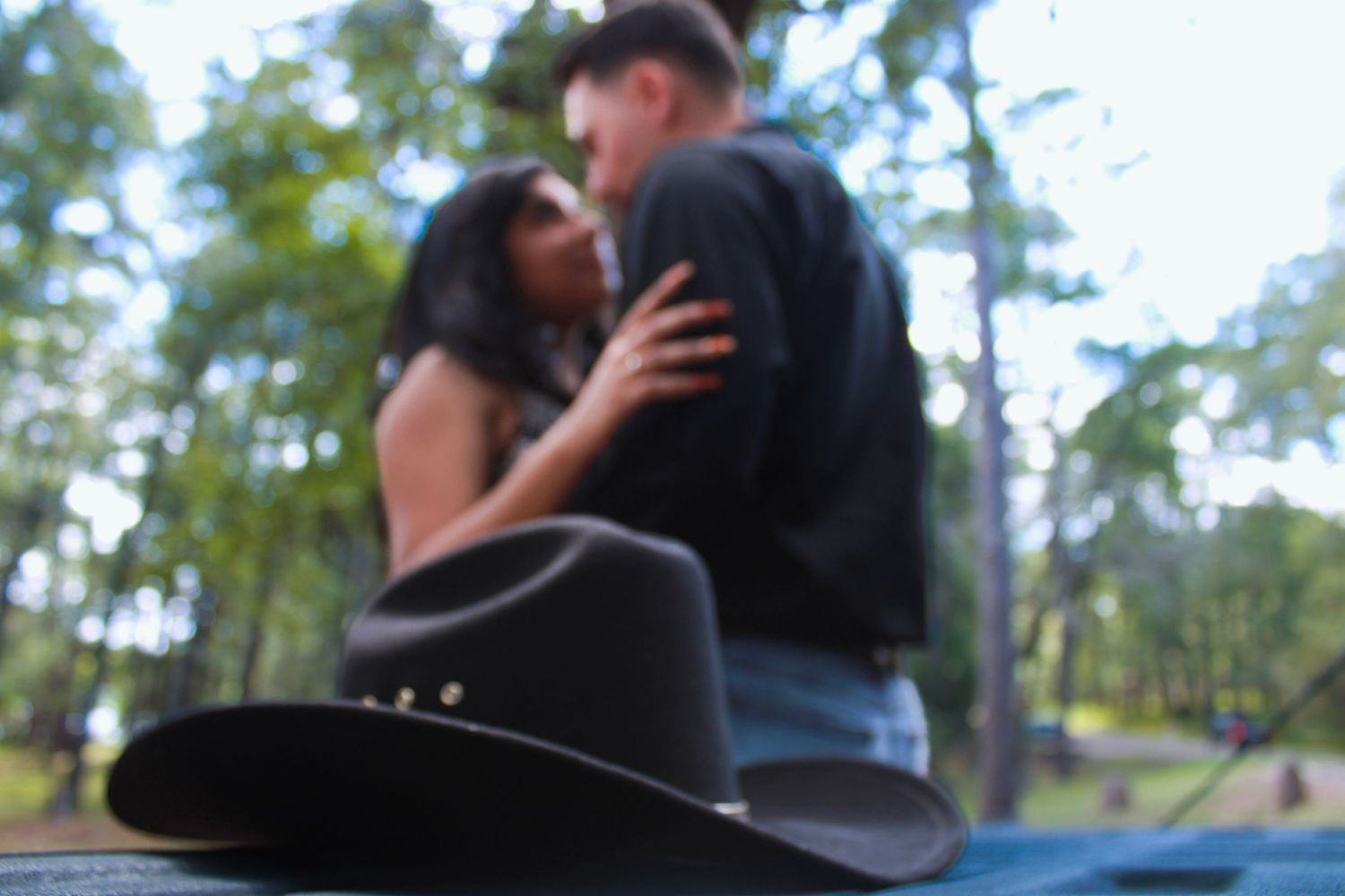 A couple shares a romantic moment near a black cowboy hat with trees in the background.