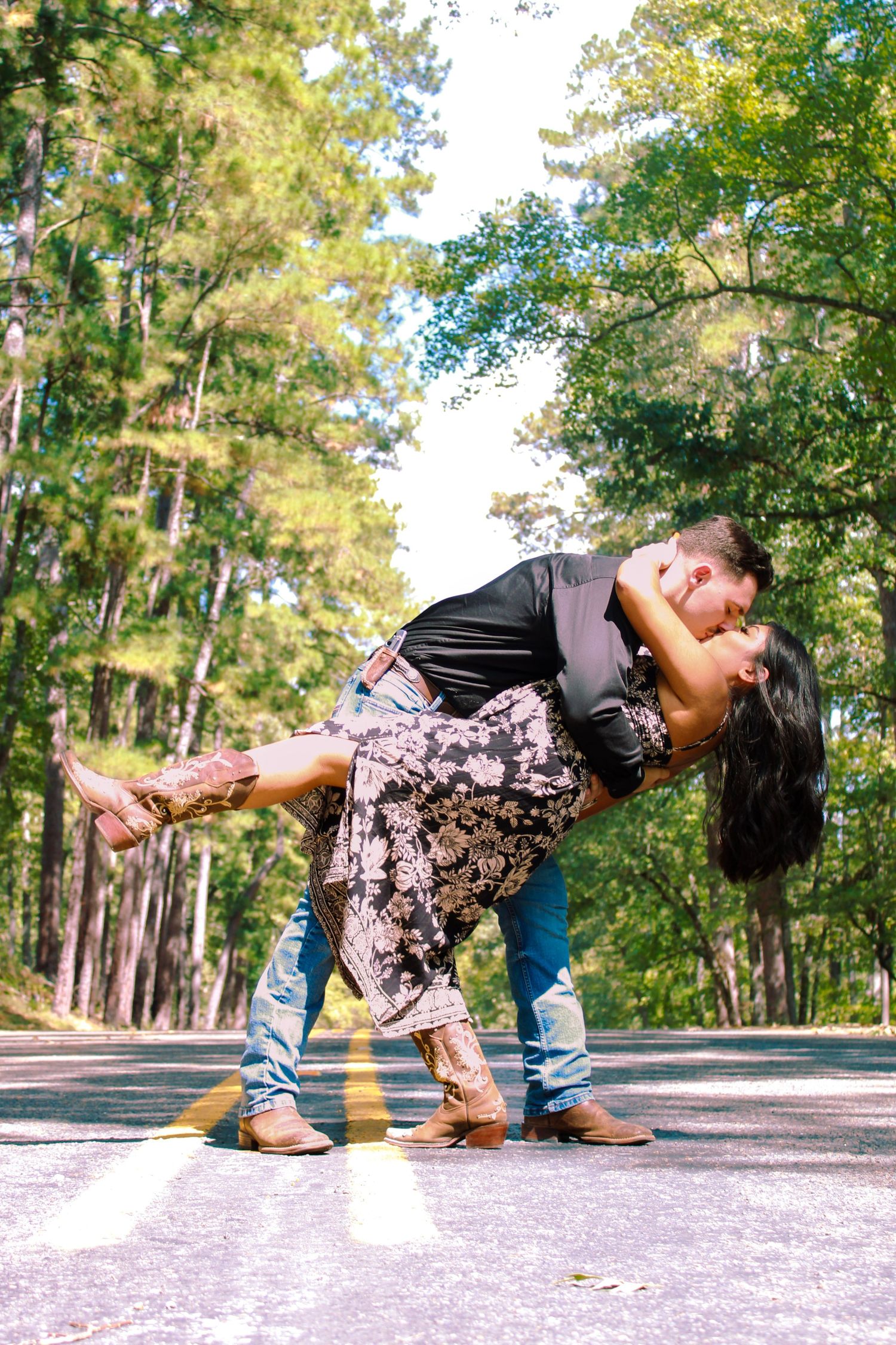 A couple performs a dramatic dance dip pose on a paved path surrounded by pine trees.