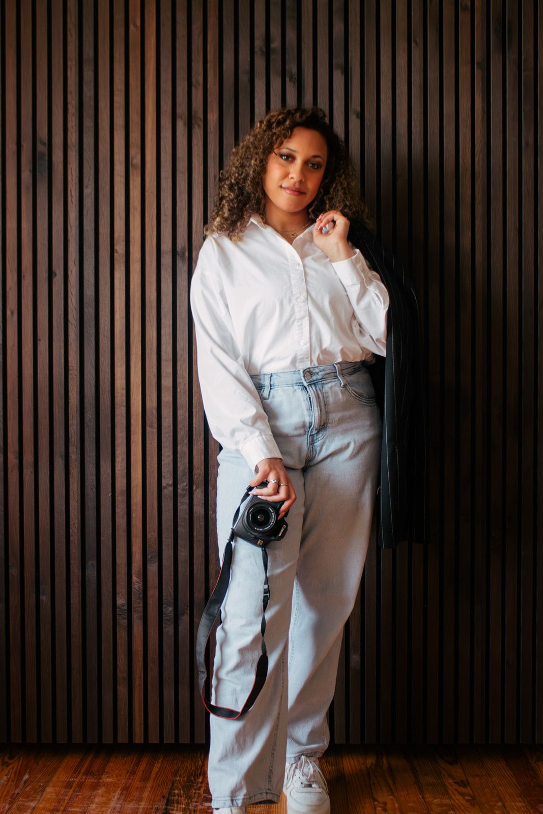 A photographer in casual white shirt and jeans poses with camera against wooden wall.