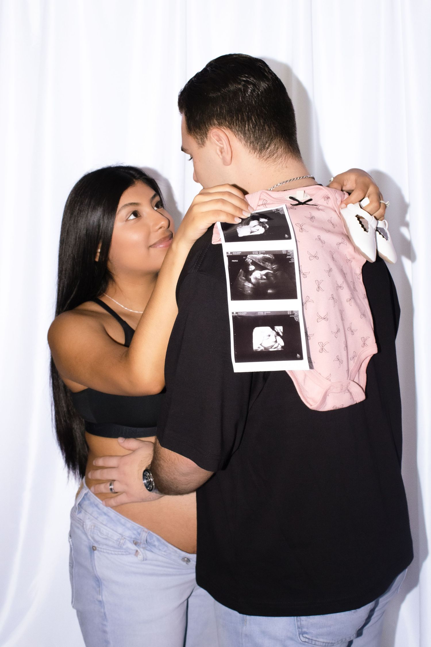 A couple shares an intimate pregnancy announcement moment while holding ultrasound photos and a pink baby outfit.