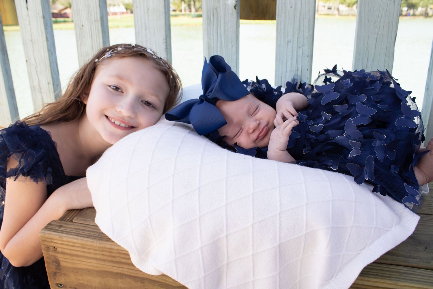 Child in navy dress lying next to sleeping baby on bench.