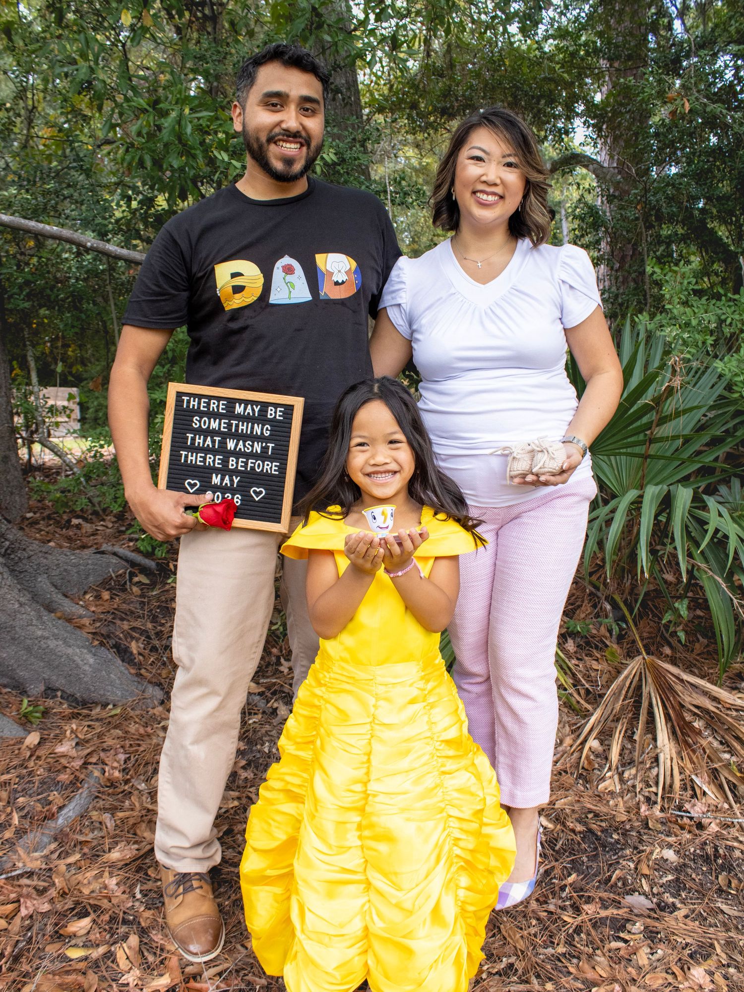 Family of three poses outdoors with a letterboard and yellow dress against a natural backdrop.