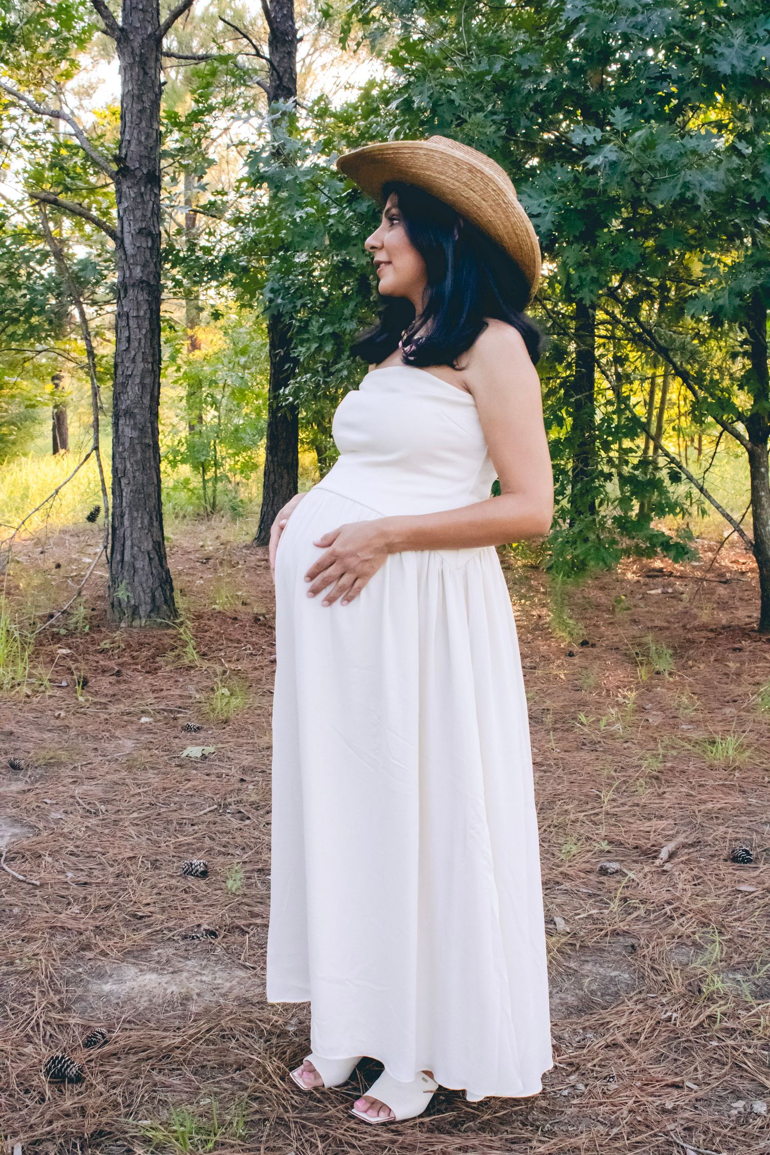 A strapless white maxi dress and straw hat create an ethereal maternity photo in a wooded setting.