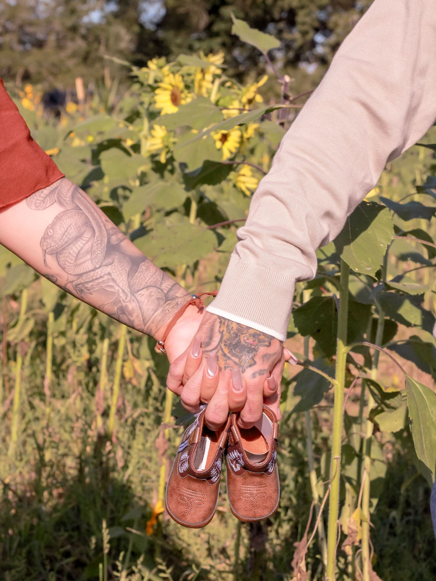 Hands holding small brown baby shoes against a backdrop of yellow sunflowers in a garden.