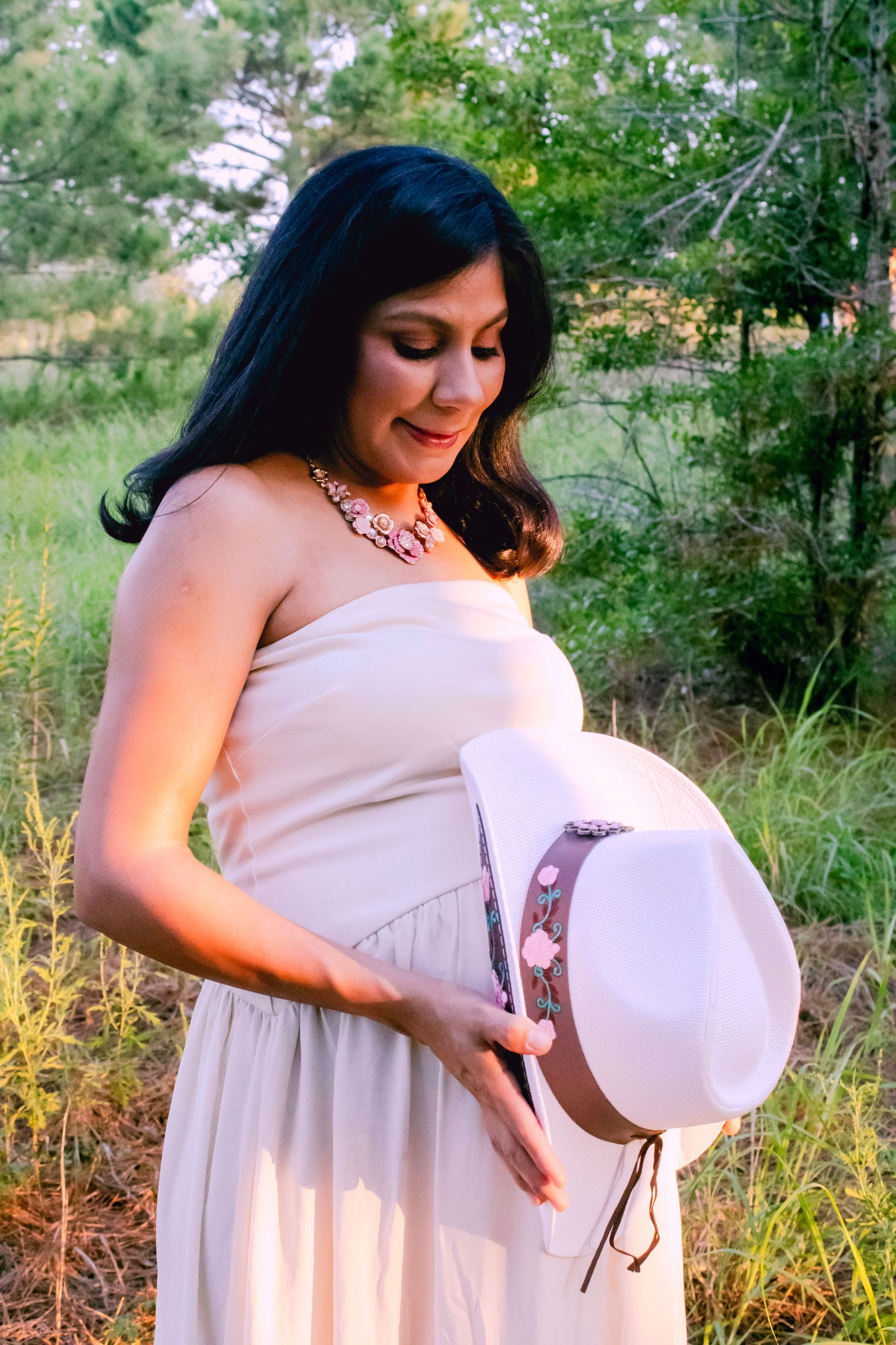 A flowing cream dress and straw hat create an elegant maternity portrait in nature.