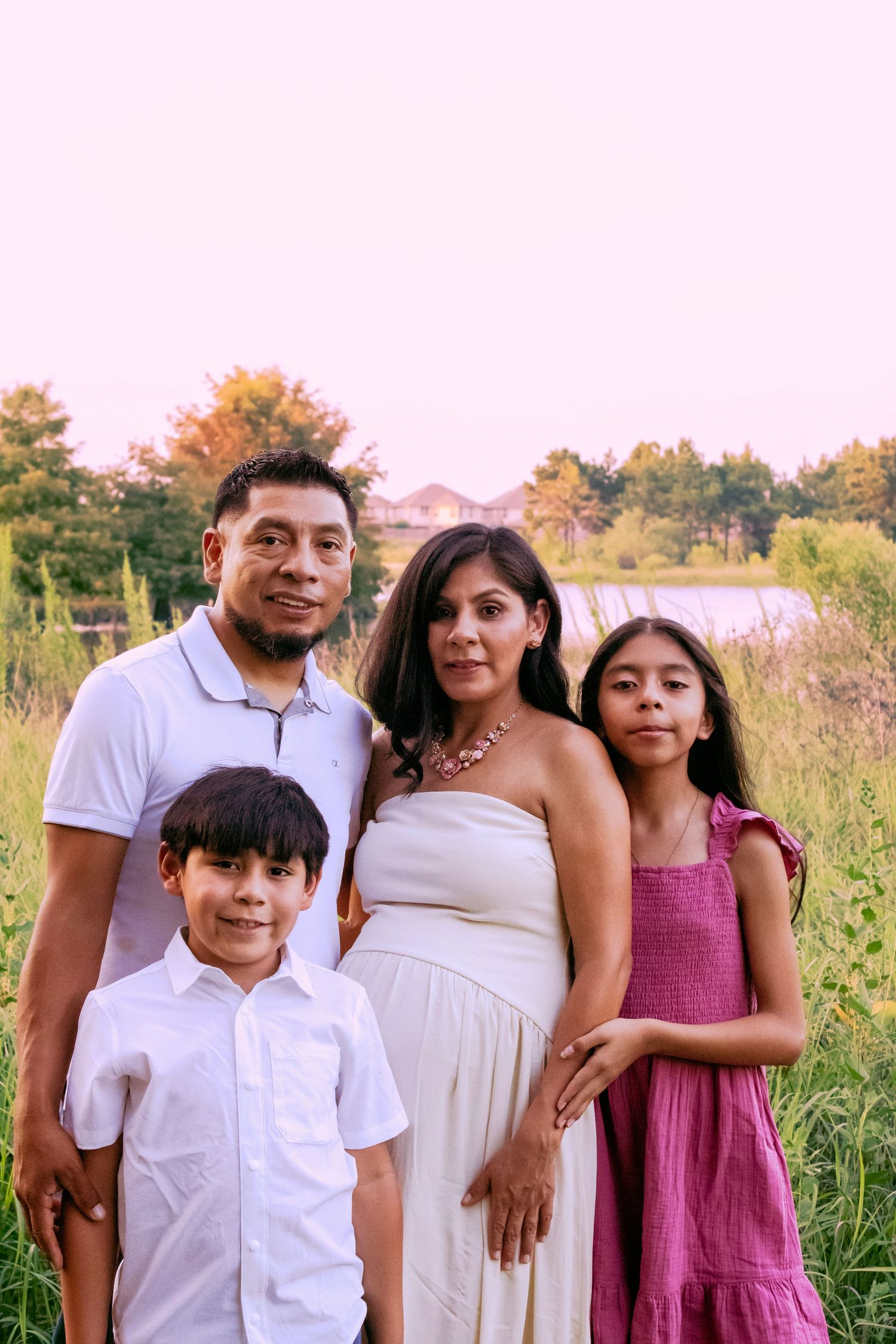 Family portrait in coordinated white and pink outfits standing in a summer field.