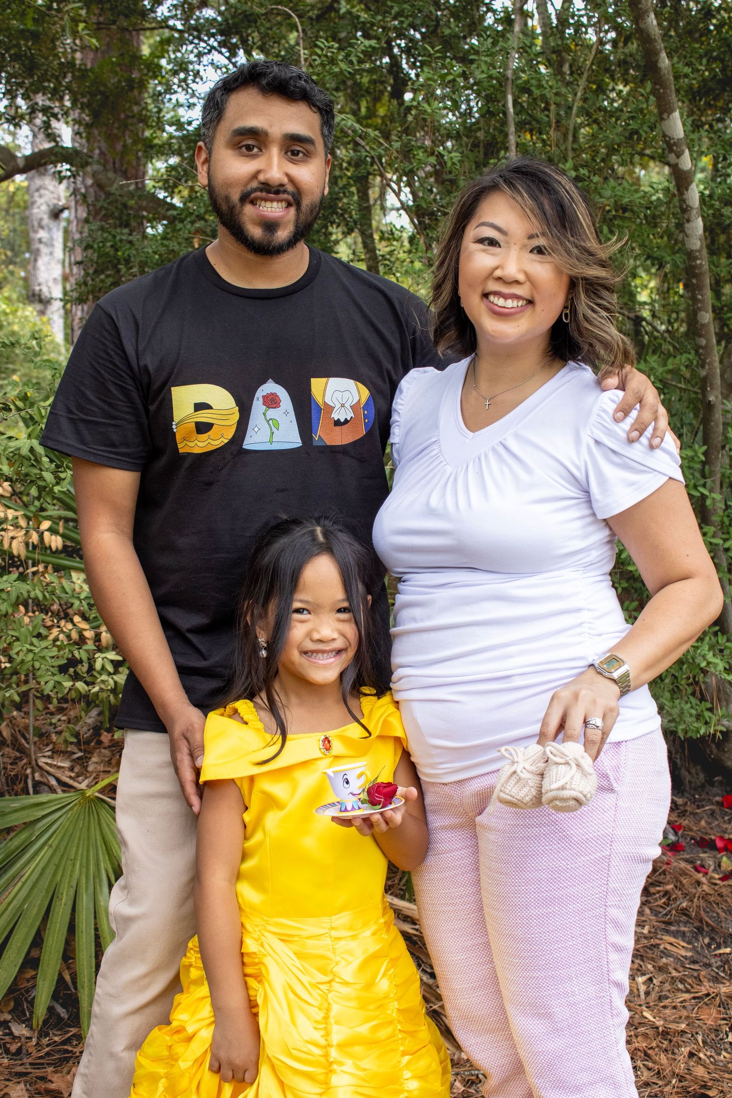 Family poses together outdoors with dad wearing 'DAD' shirt and child in yellow dress.