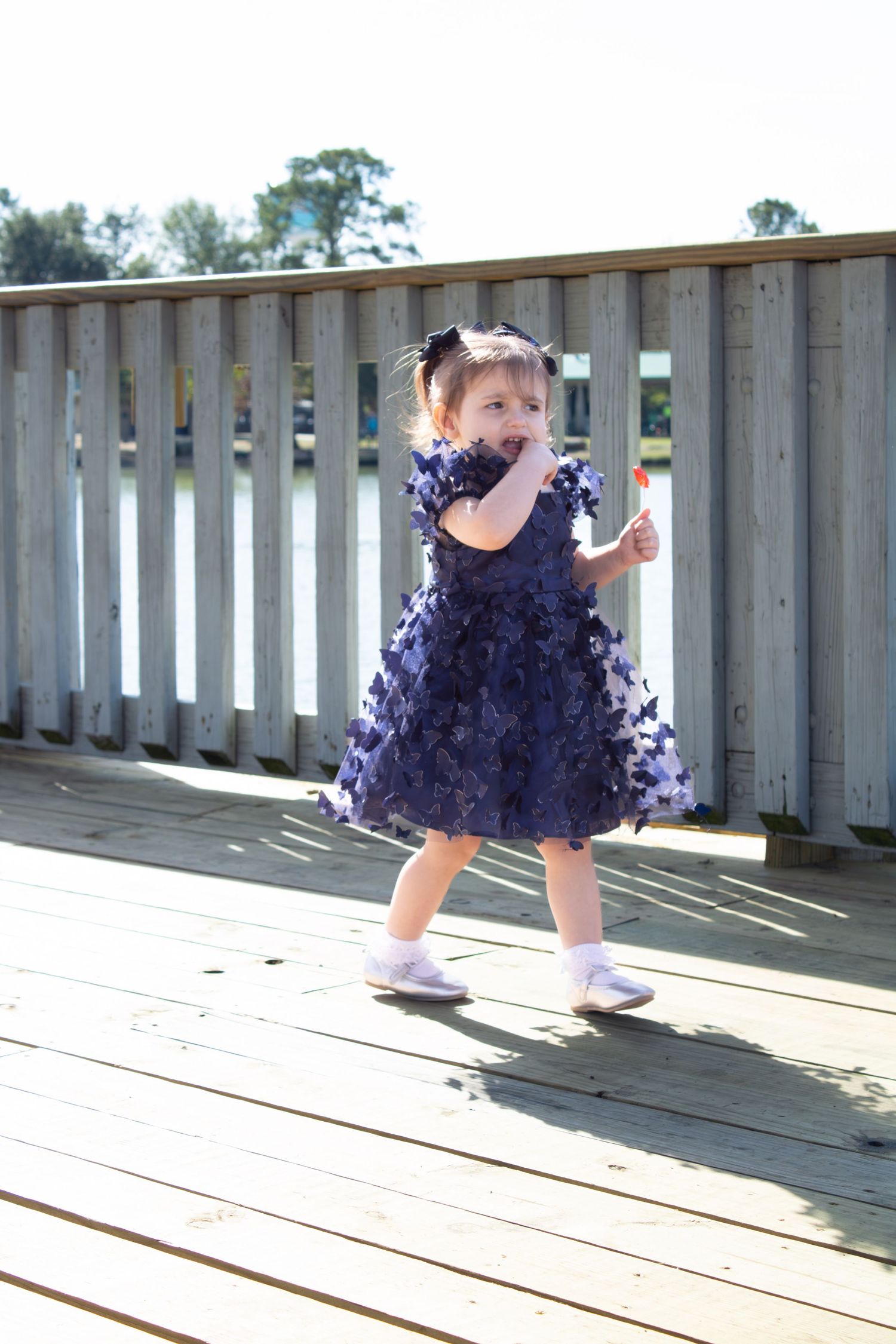 Young child in a navy blue tulle dress twirls and plays on a wooden deck in sunlight.